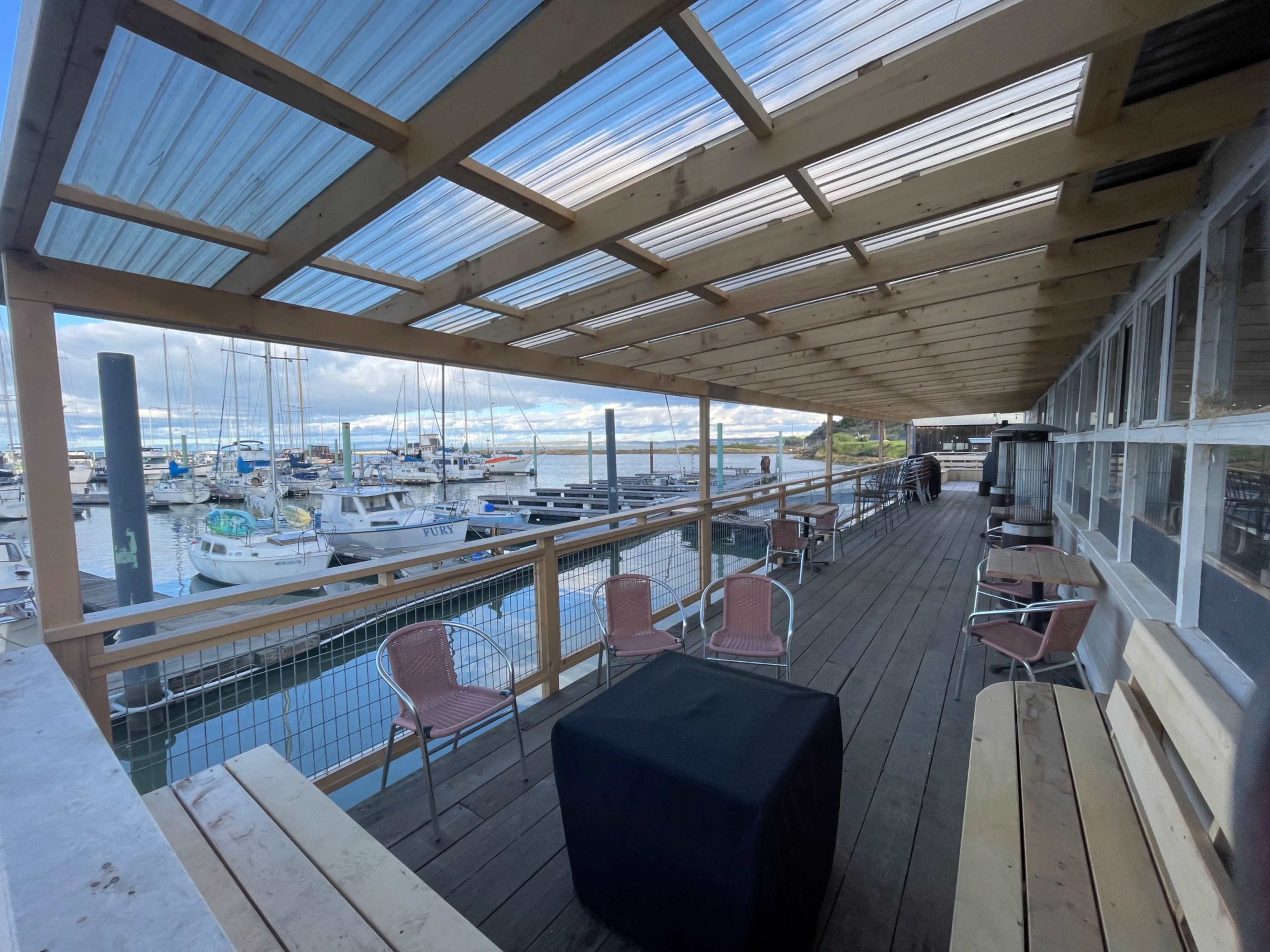 A covered wooden deck overlooking a marina filled with boats, featuring outdoor seating arrangements and a table.