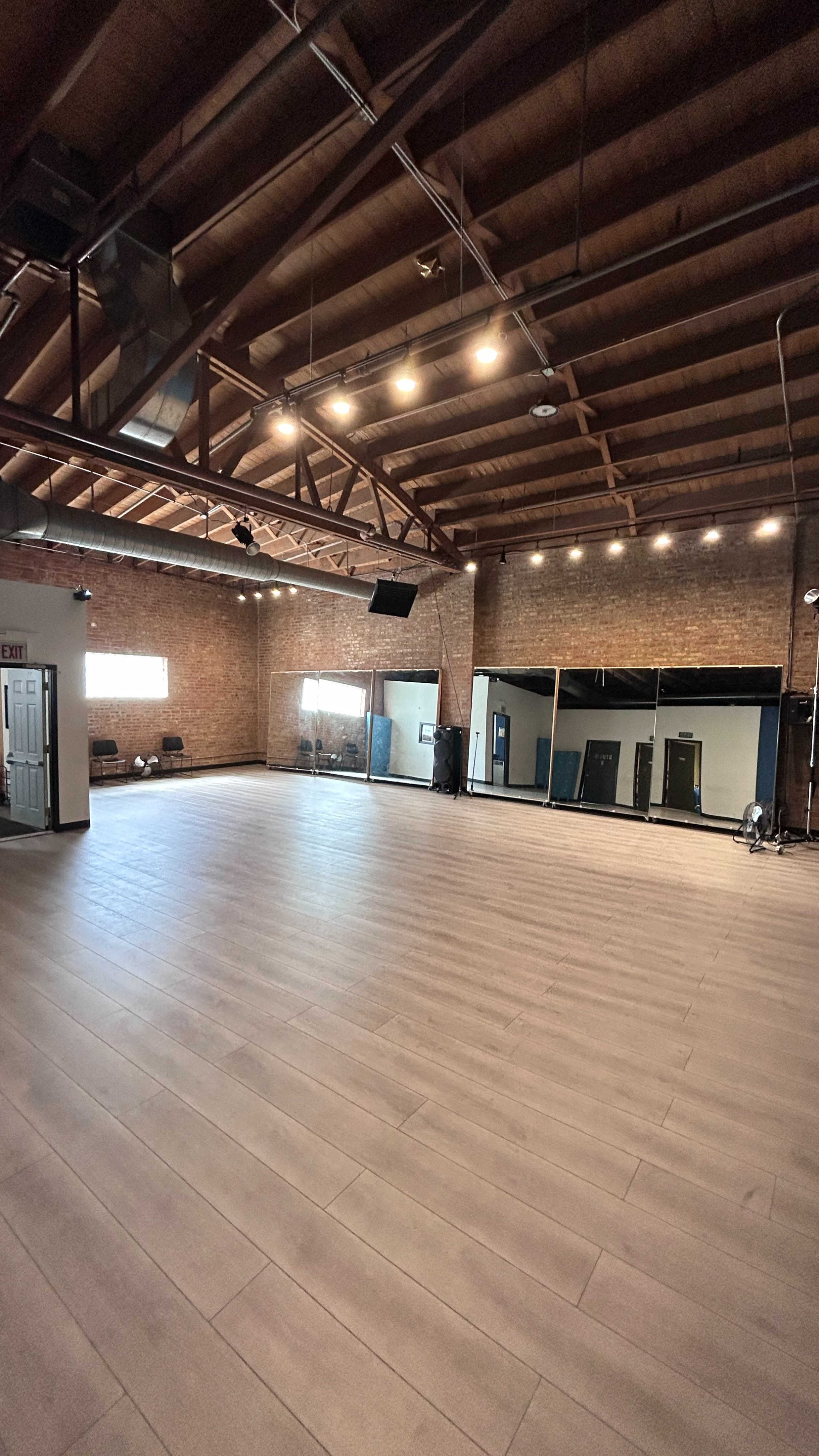 The image shows an empty studio space with wooden beams, a large smooth floor, and exposed brick walls.