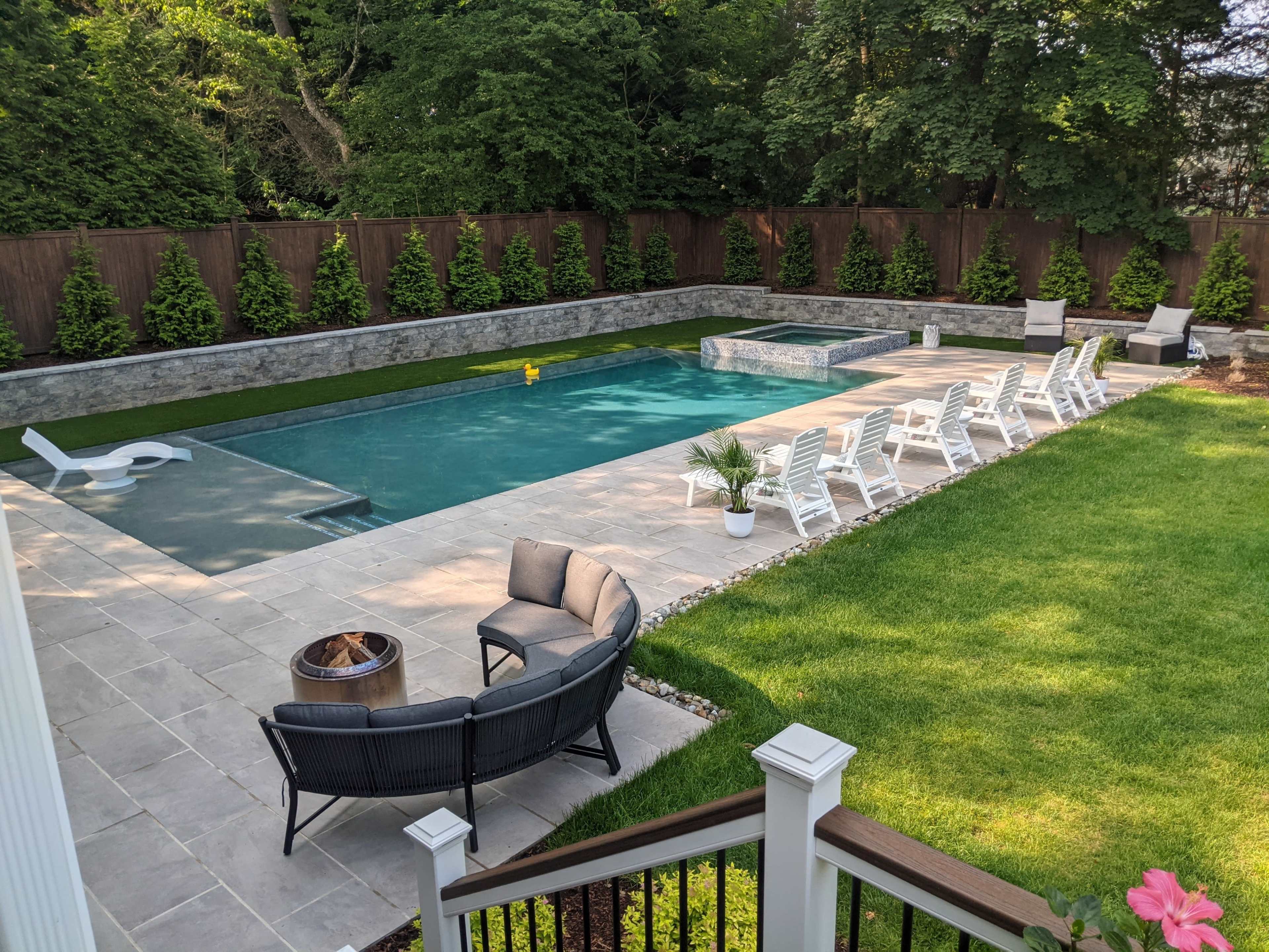A backyard with a rectangular swimming pool surrounded by lounge chairs, stone paths, and landscaped greenery.