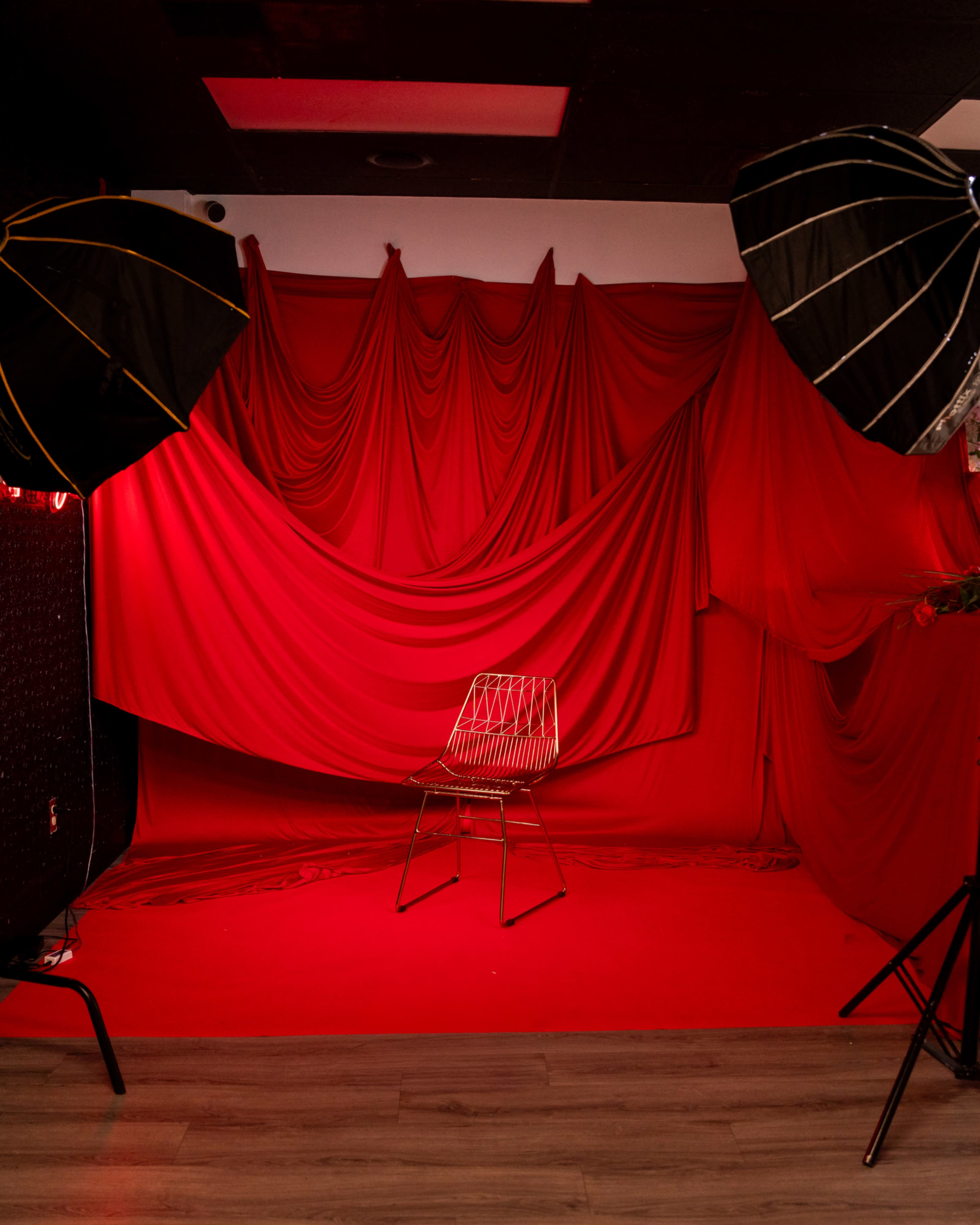 The image shows a photography studio with a red backdrop and lighting, featuring an empty wire chair at the center.