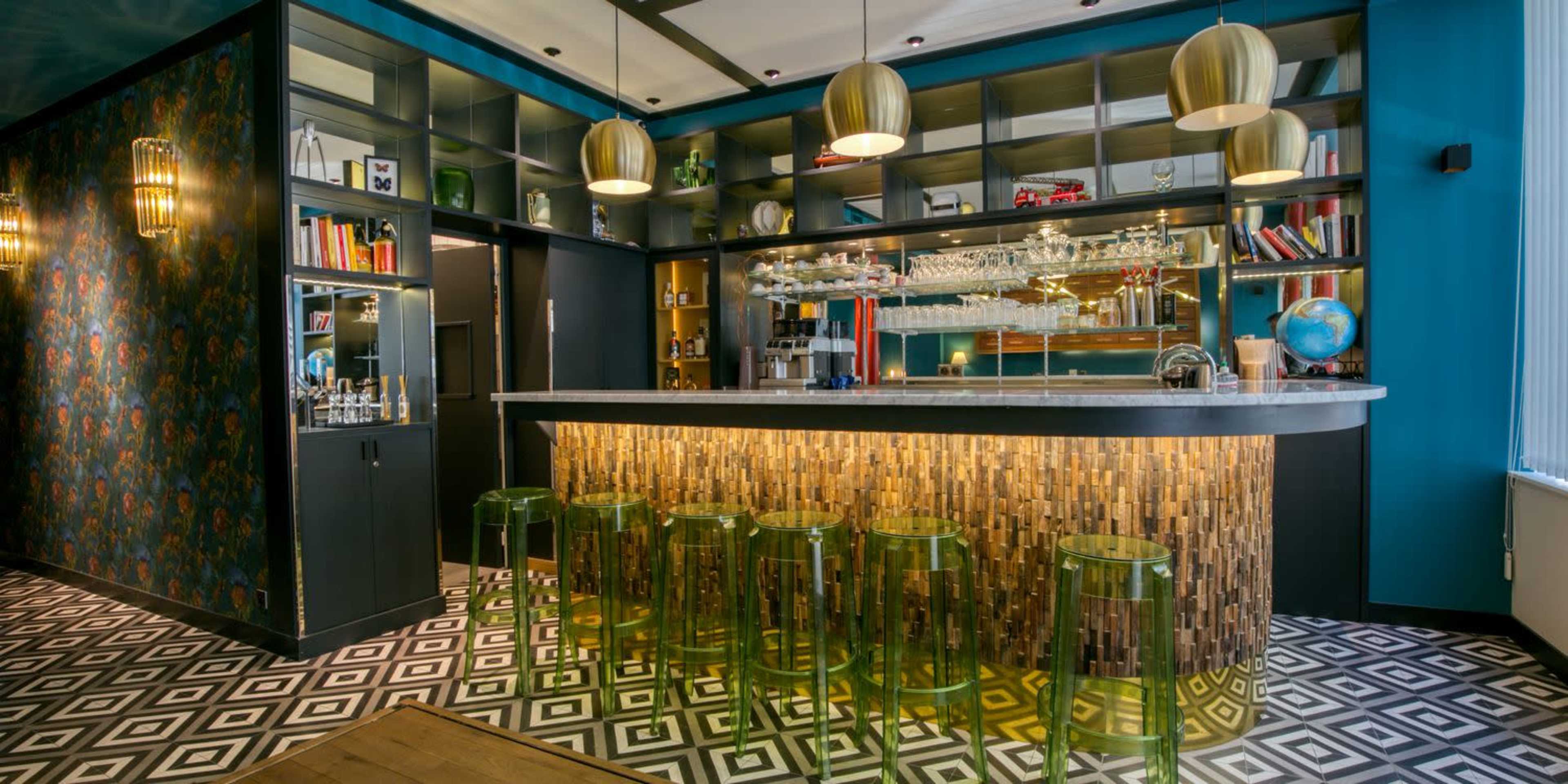 A modern bar with a curved countertop, illuminated by gold pendant lights, surrounded by sleek green stools and a backdrop of shelves filled with glassware and books.