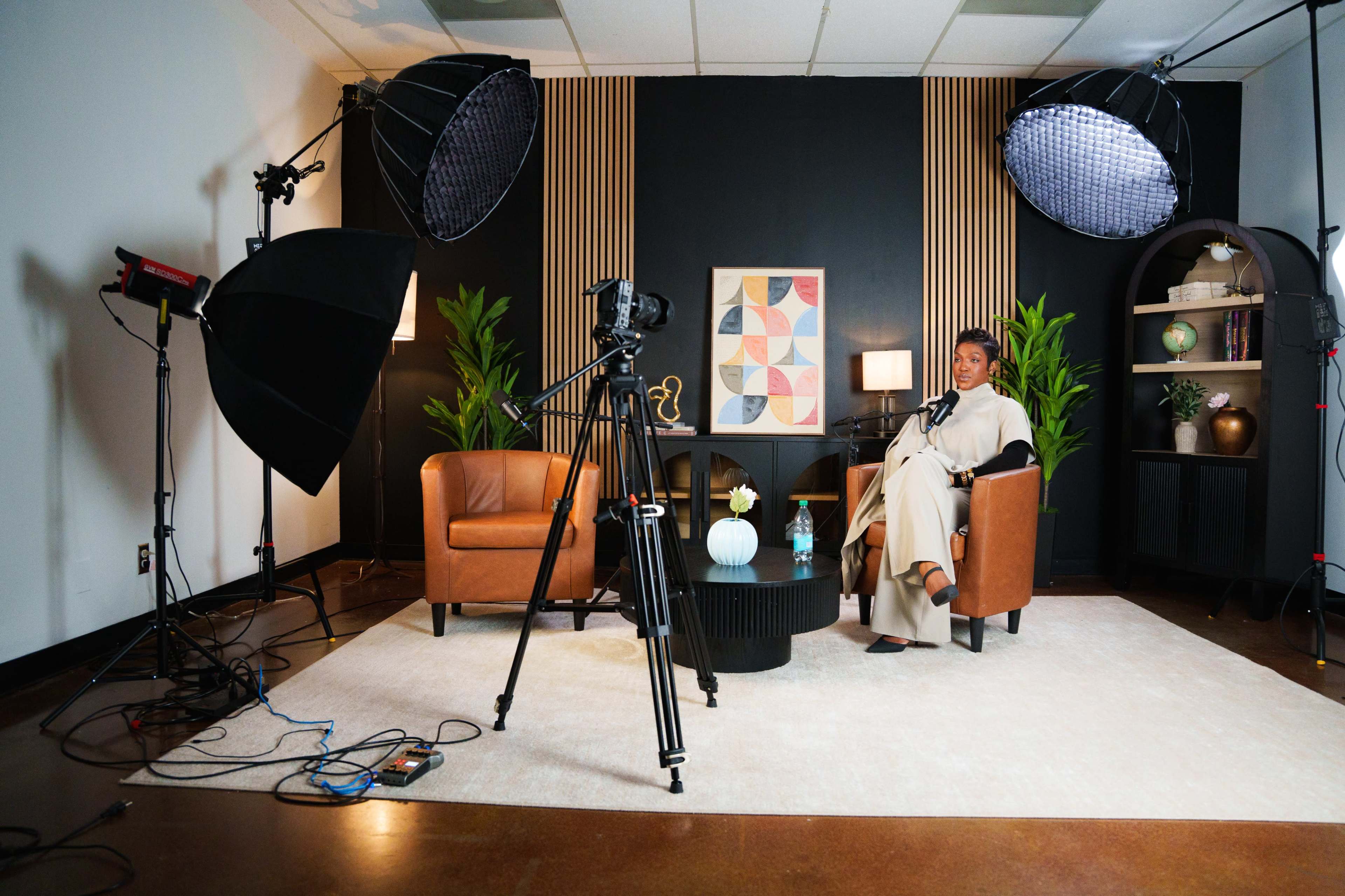 A well-lit studio setting features a woman sitting on a chair next to a coffee table, surrounded by cameras and studio lighting equipment.
