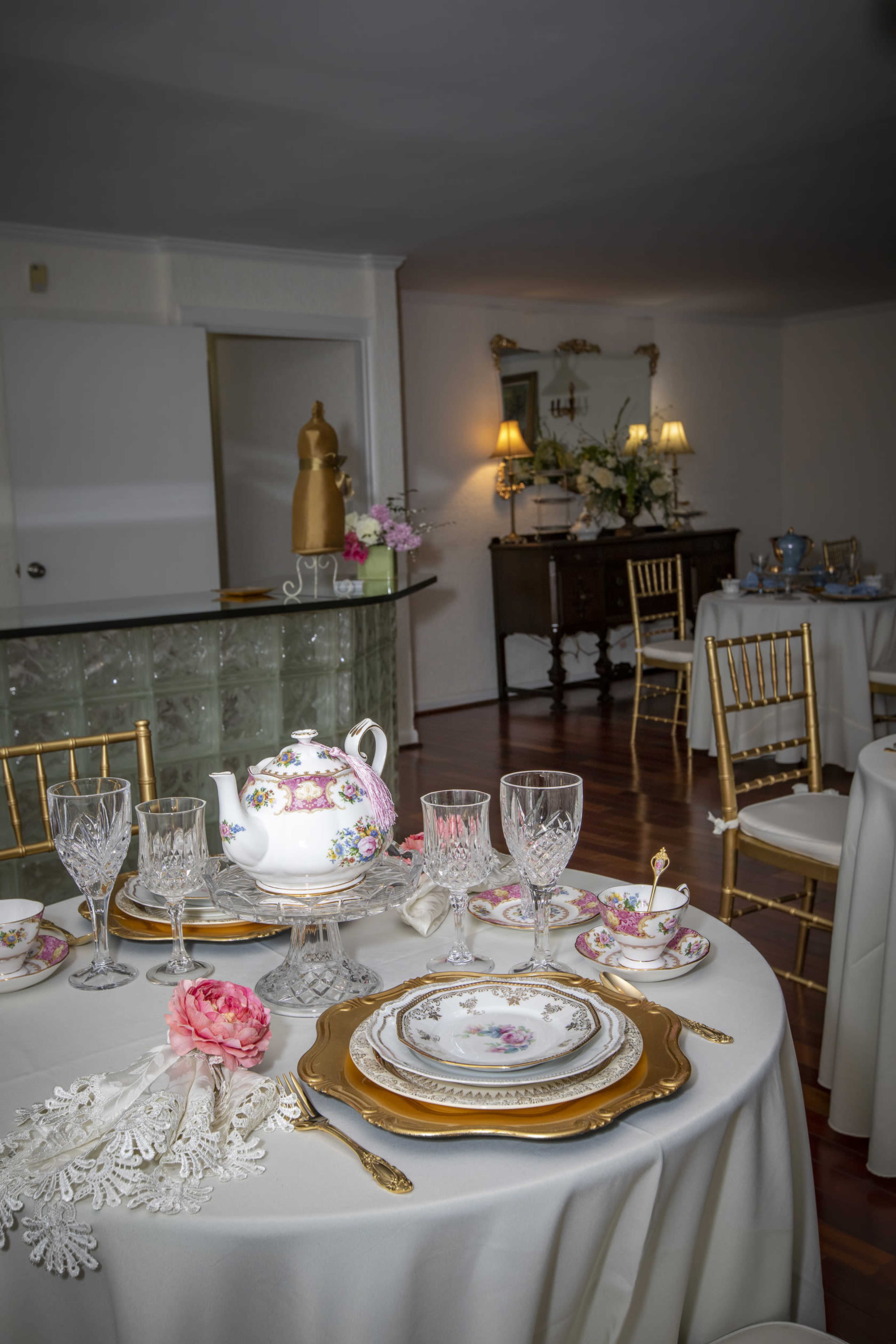 A neatly set dining table features fine china, crystal glassware, and lace napkins, surrounded by elegantly arranged chairs in a softly lit room.