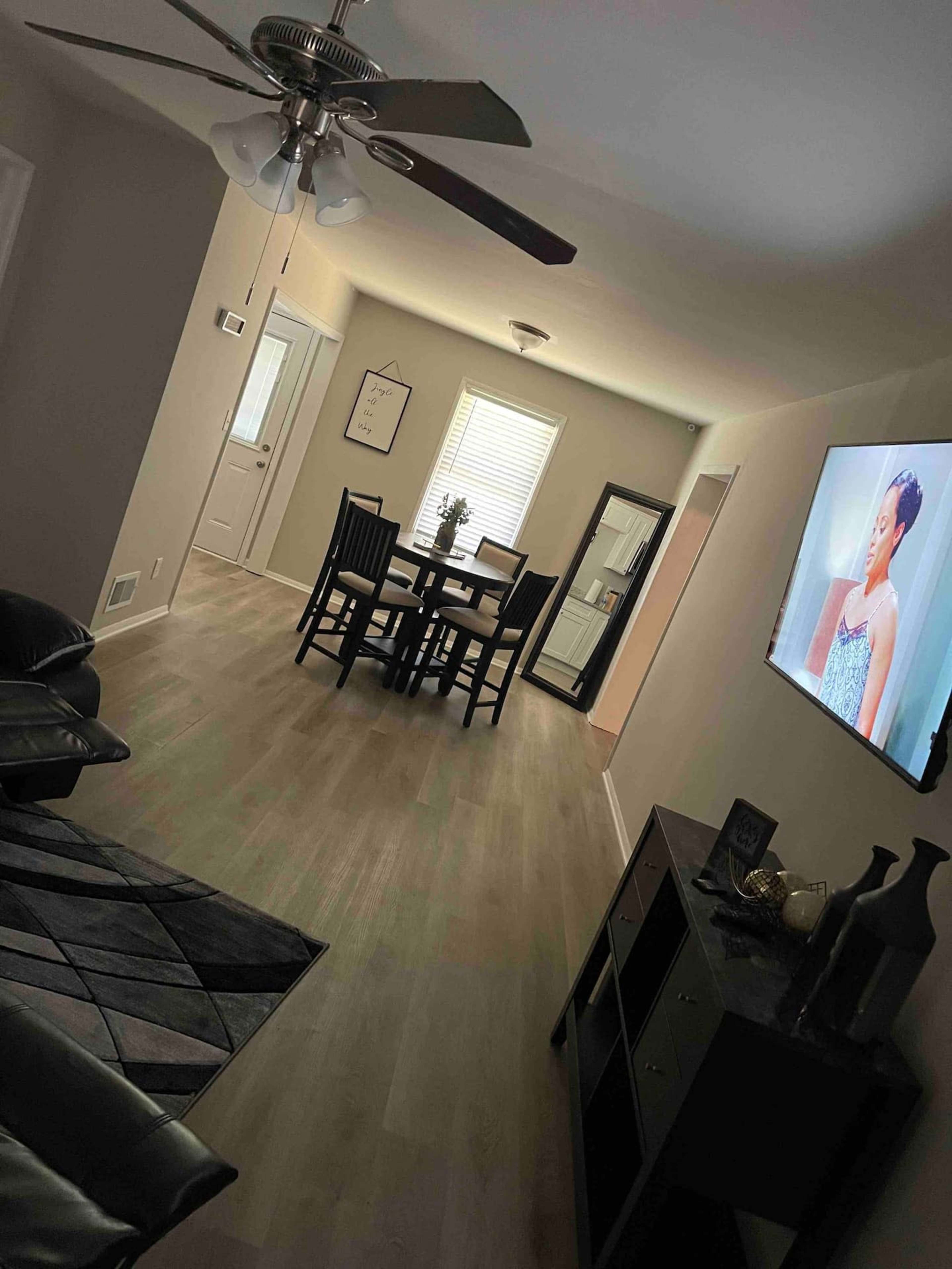 A living room with a fan, a black sofa, a dining area with a table and chairs, and a television displaying a woman.