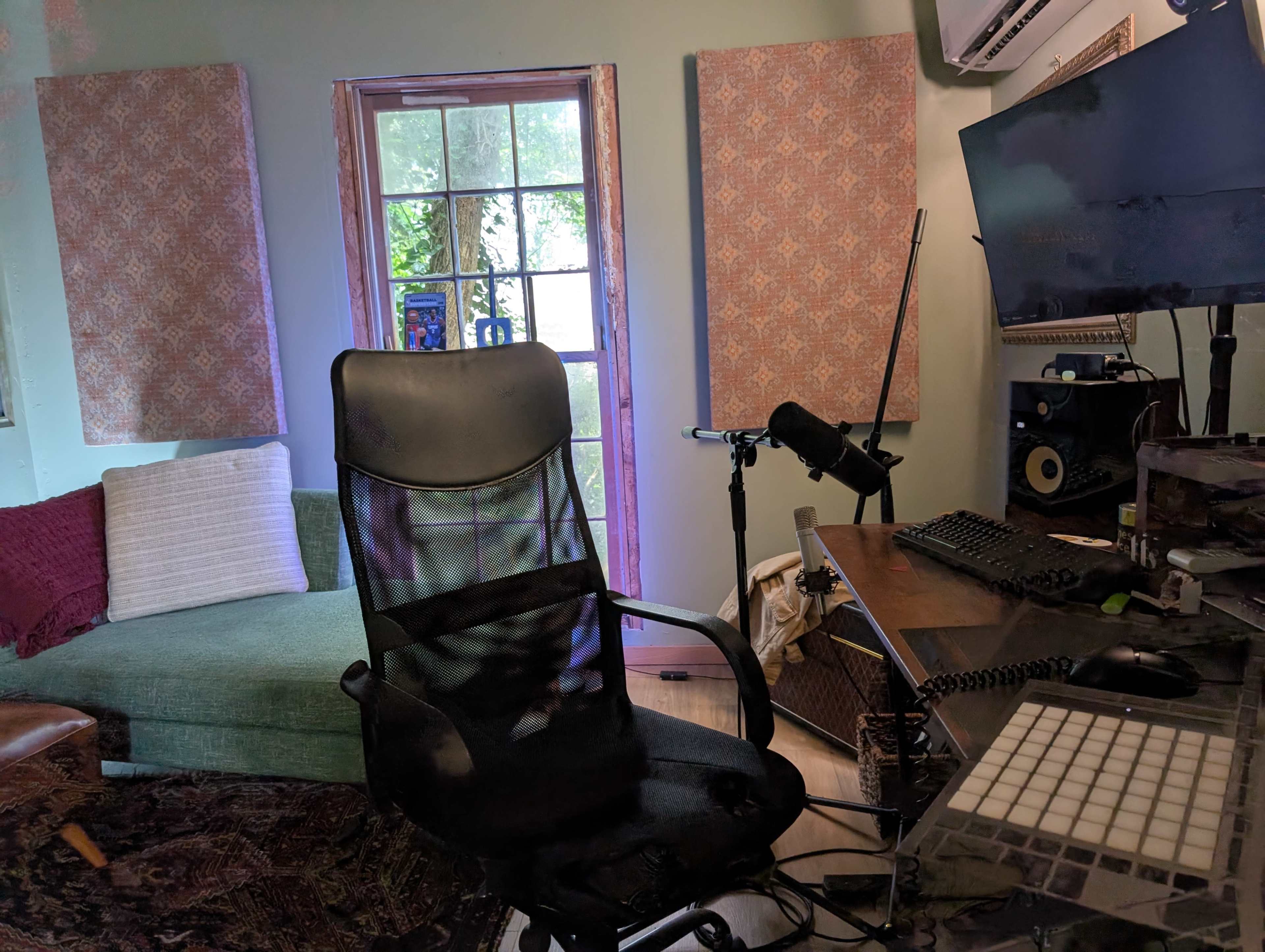 The image shows a small home office with a desk, computer monitor, microphone, and a green sofa near a window with fabric-covered panels.