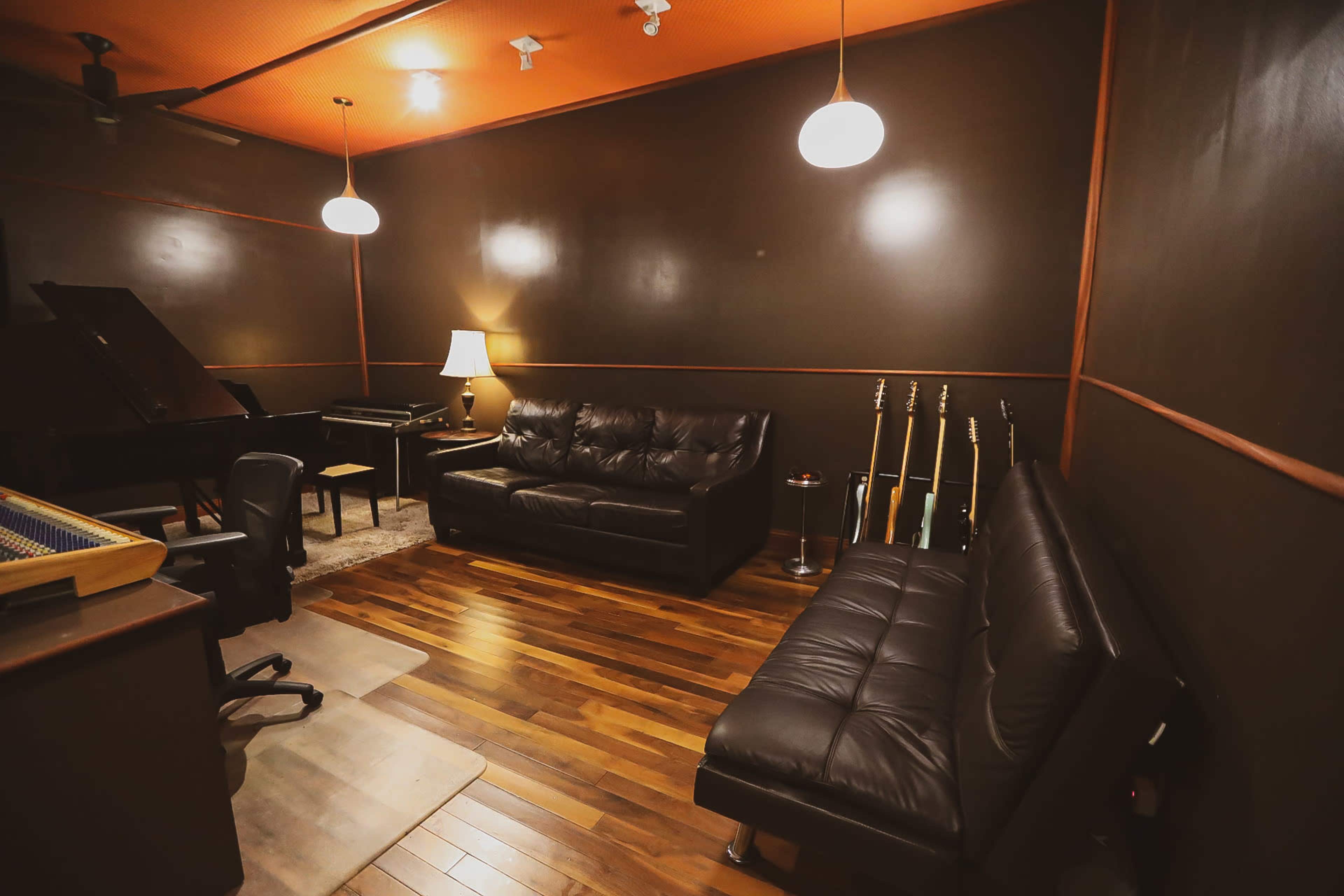 The image depicts a dimly lit music studio featuring a black leather couch, a grand piano, and several guitars displayed on the wall.