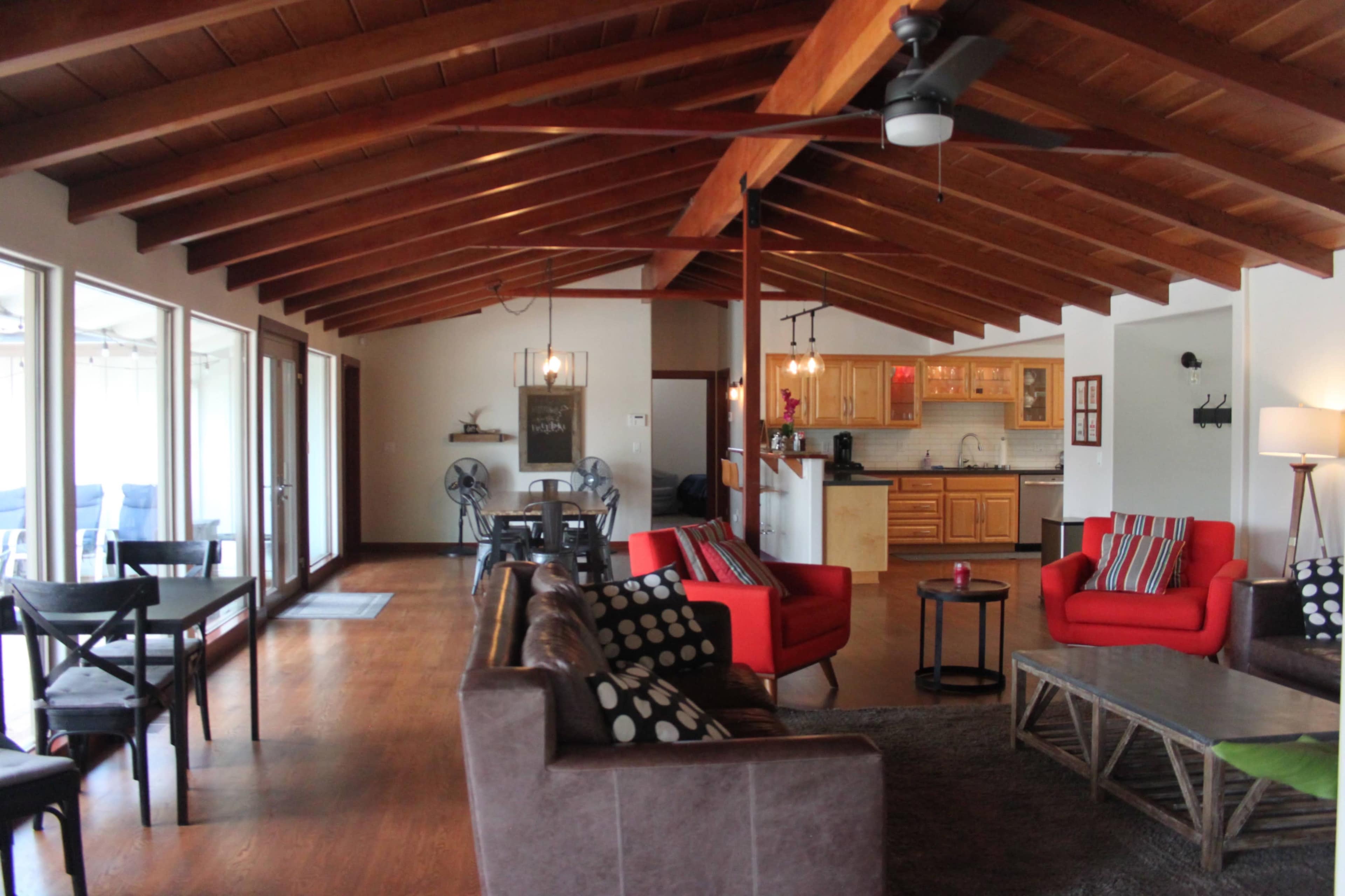 A spacious living area features a wooden ceiling, red armchairs, a sofa with patterned cushions, and large windows letting in natural light.