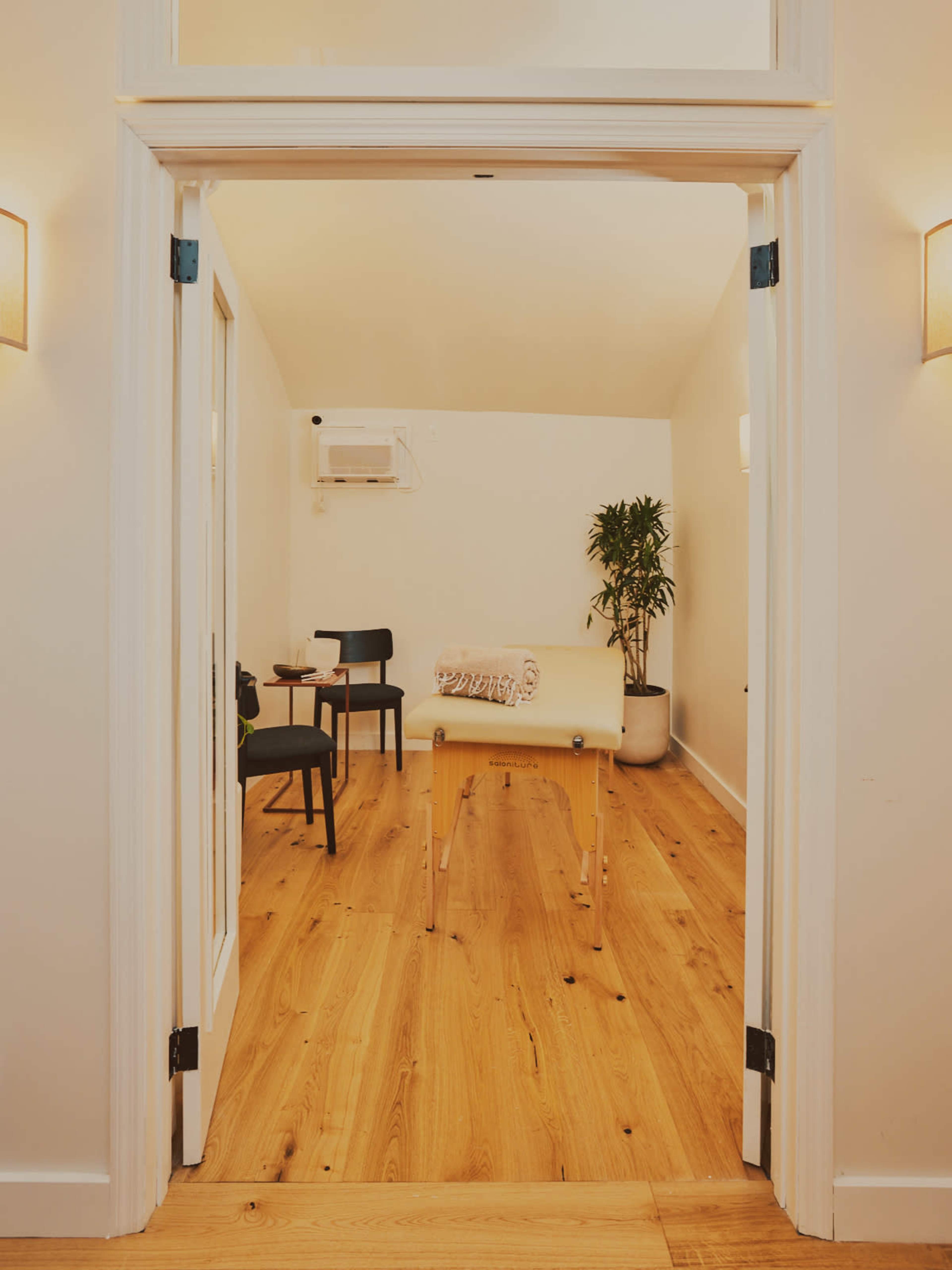 A minimalist treatment room features a massage table, a chair, and a potted plant, with wooden flooring and soft wall lighting.