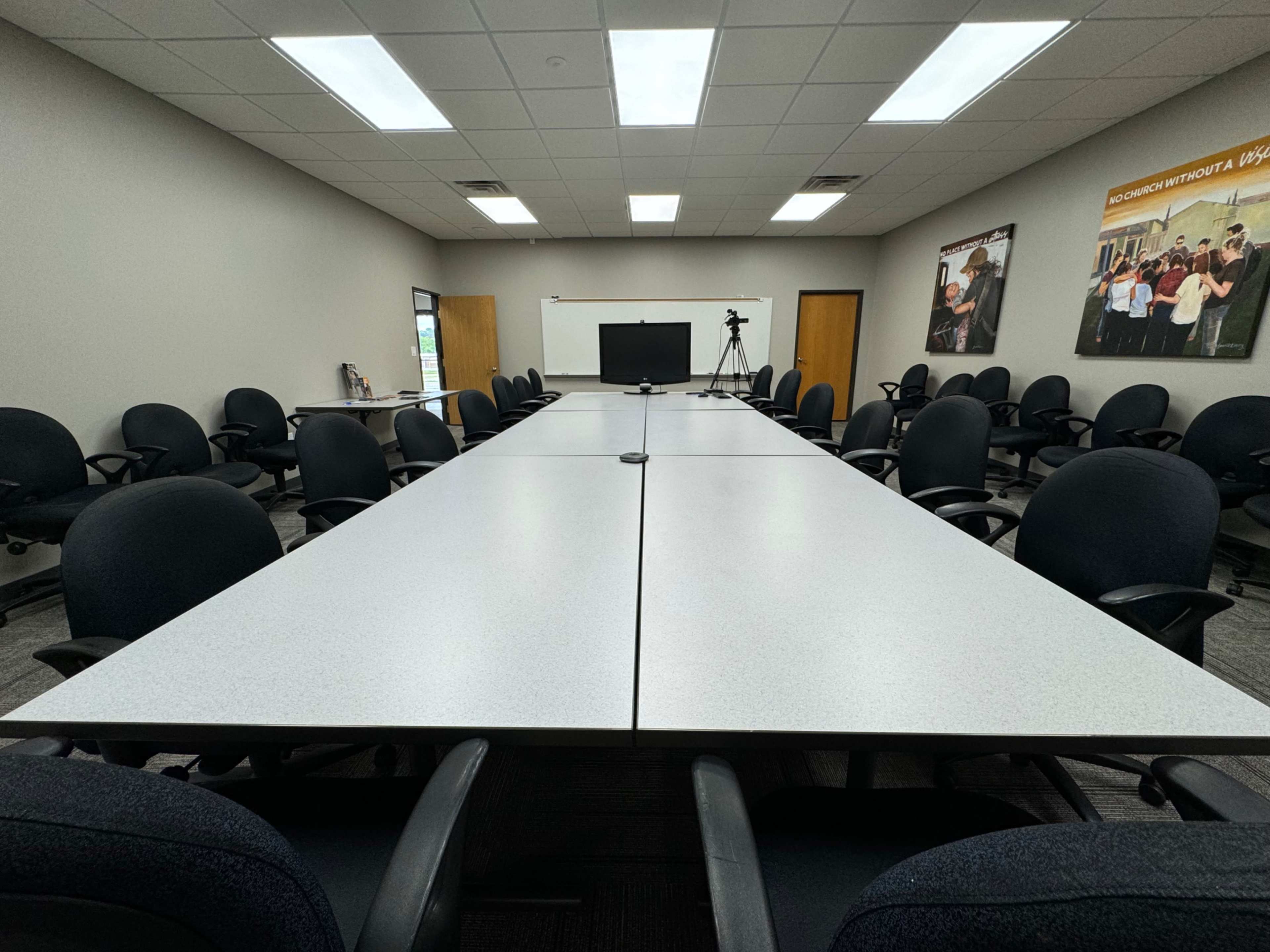 A large conference room features two long tables arranged in a U-shape with numerous chairs and a projector screen at one end.