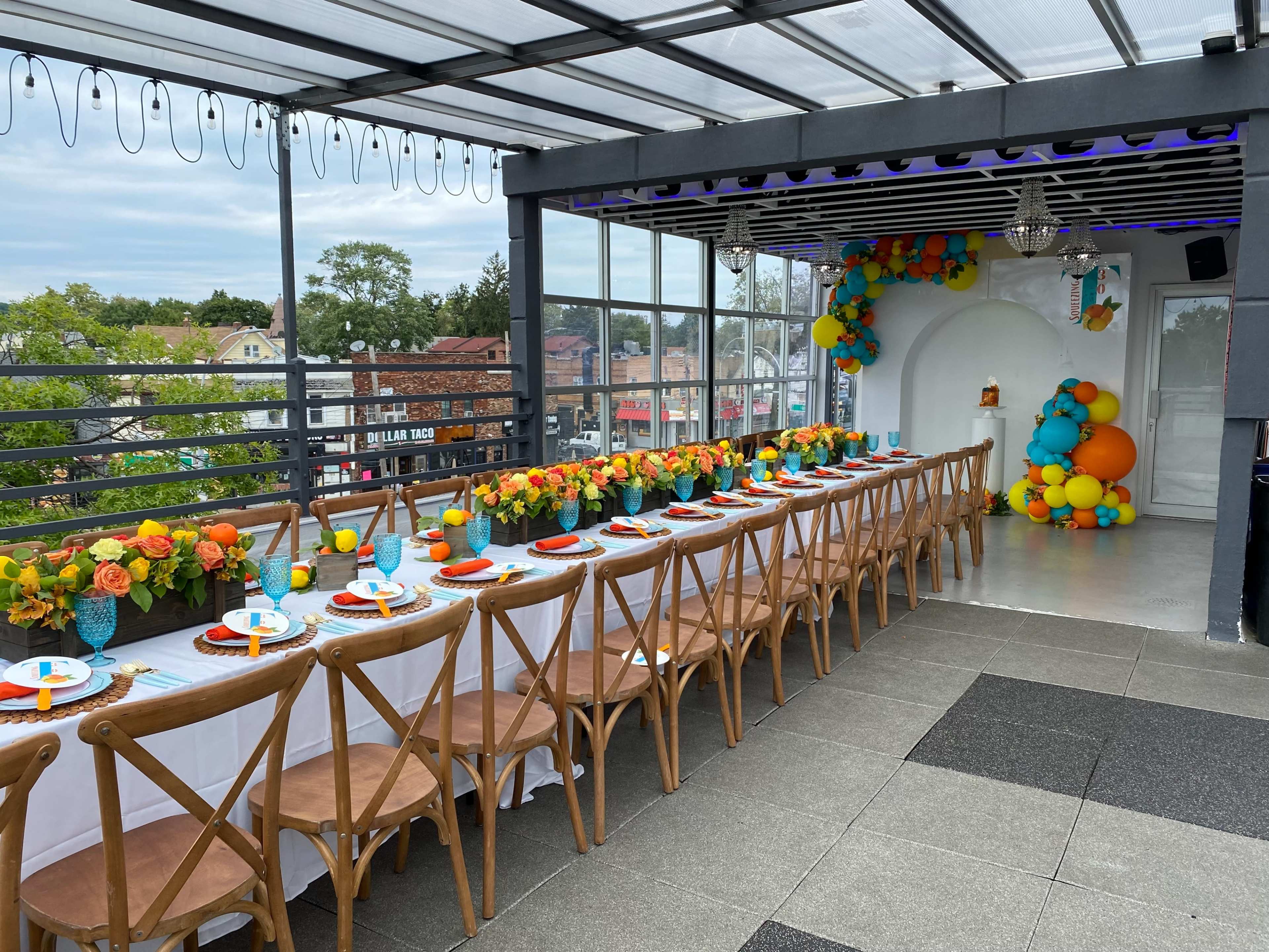 A long dining table is set up on a covered terrace, decorated with vibrant flowers and colorful balloons, overlooking a cityscape.