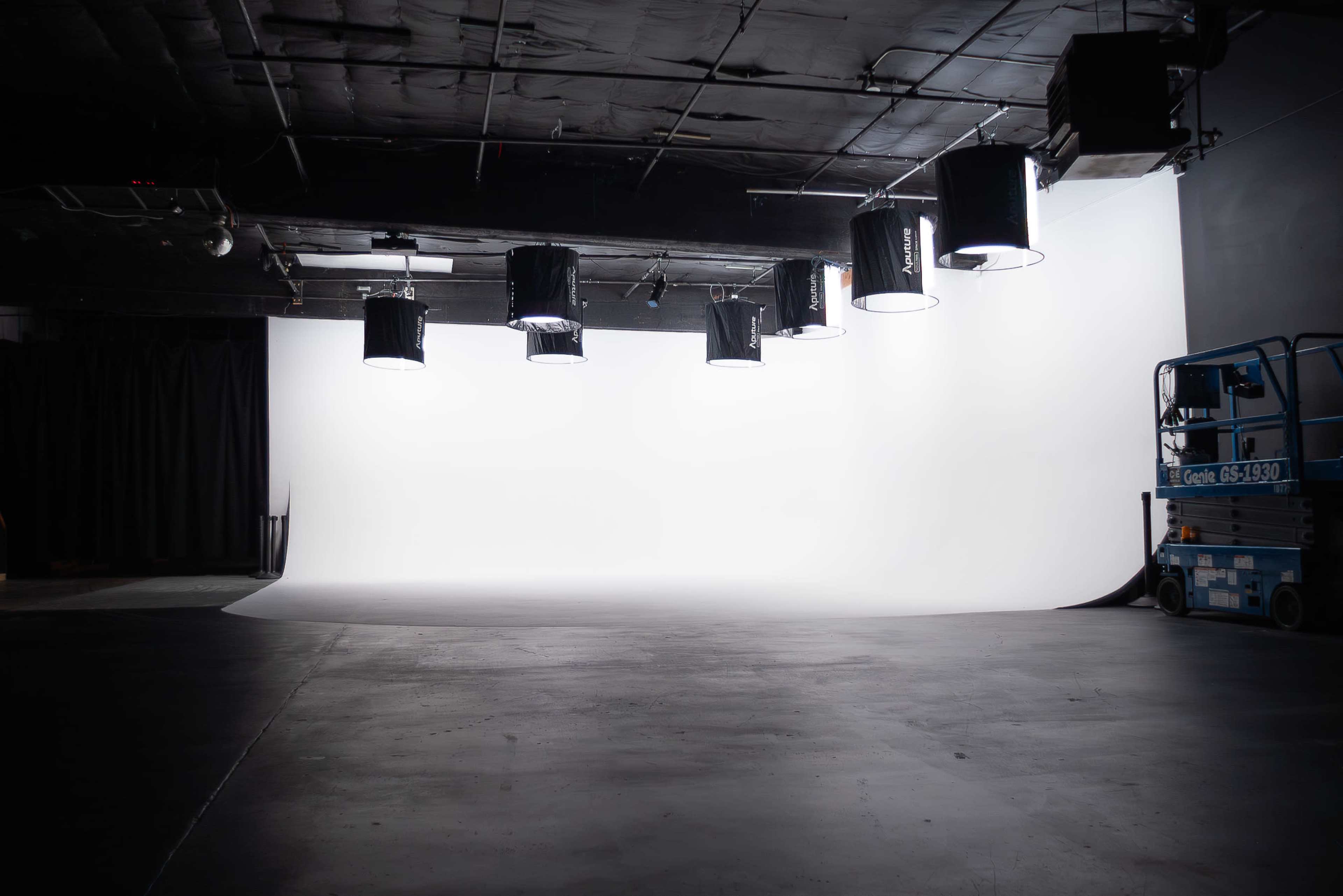 The image shows a photography studio with a blank white backdrop and multiple overhead lights.