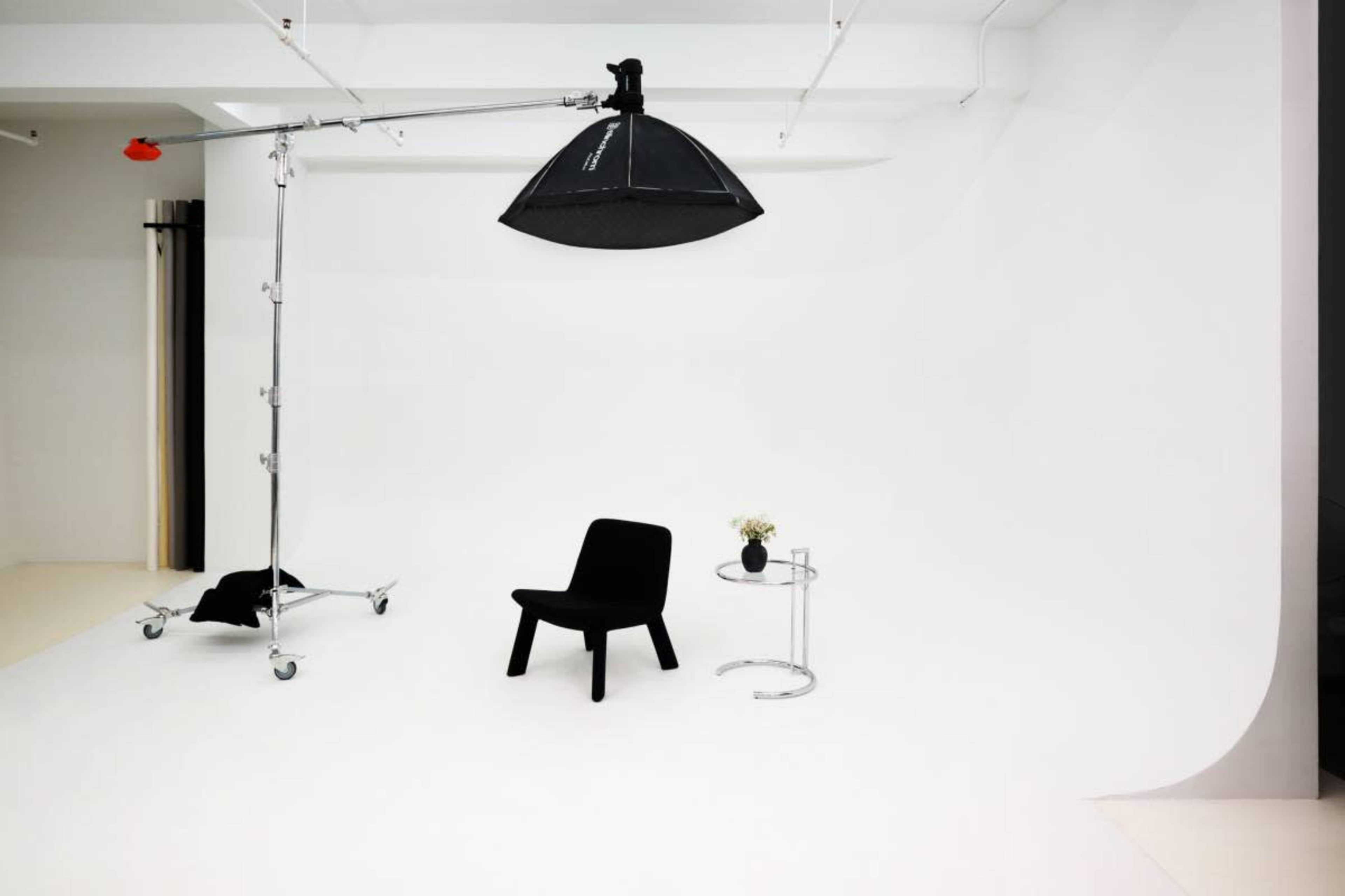 The image shows a minimalist photo studio with a white backdrop, a portable lighting setup, a black chair, and a glass side table with a flower arrangement.