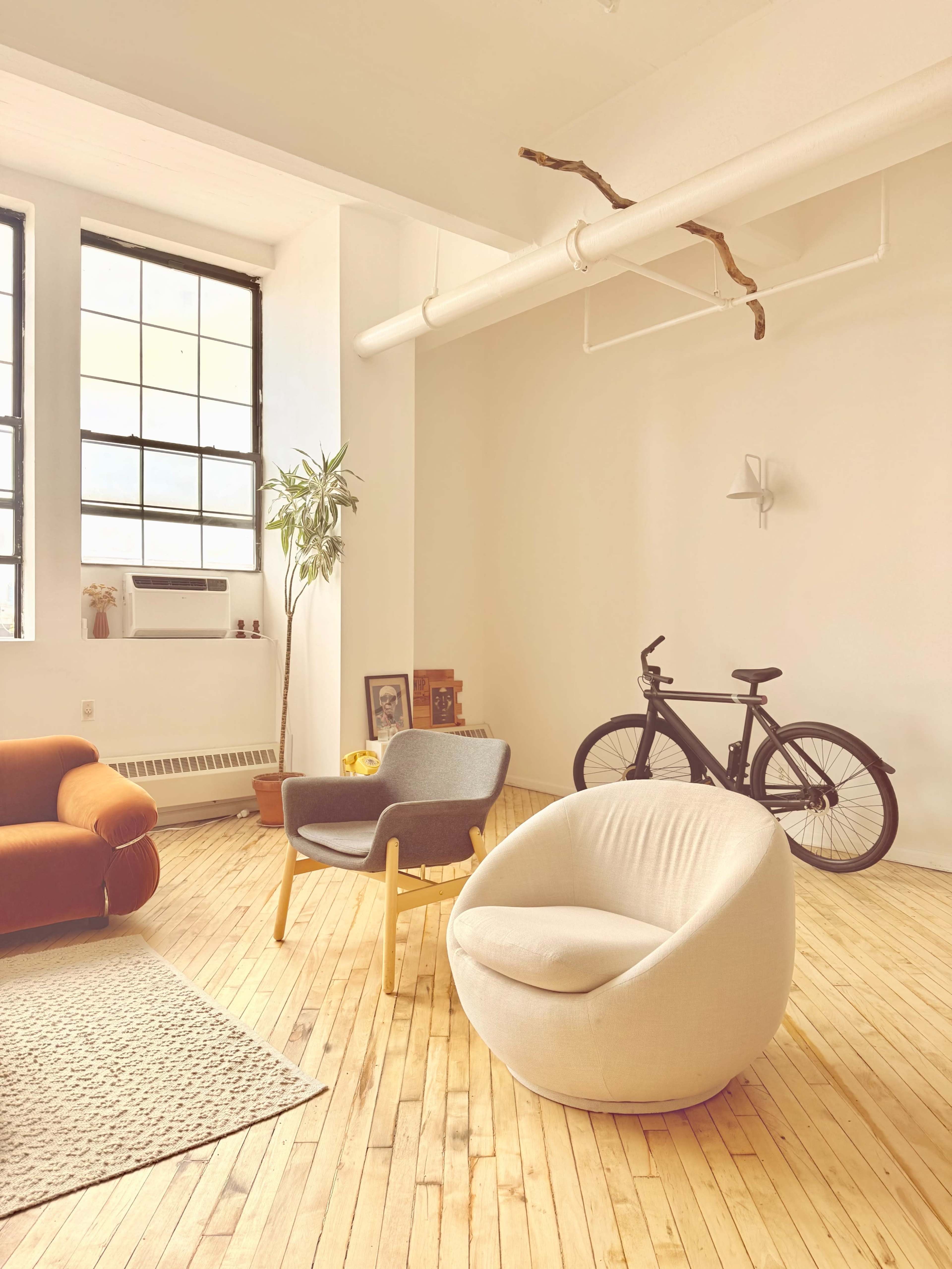 The image shows a spacious, modern living room with wooden floors, two chairs, a bicycle, and large windows letting in natural light.