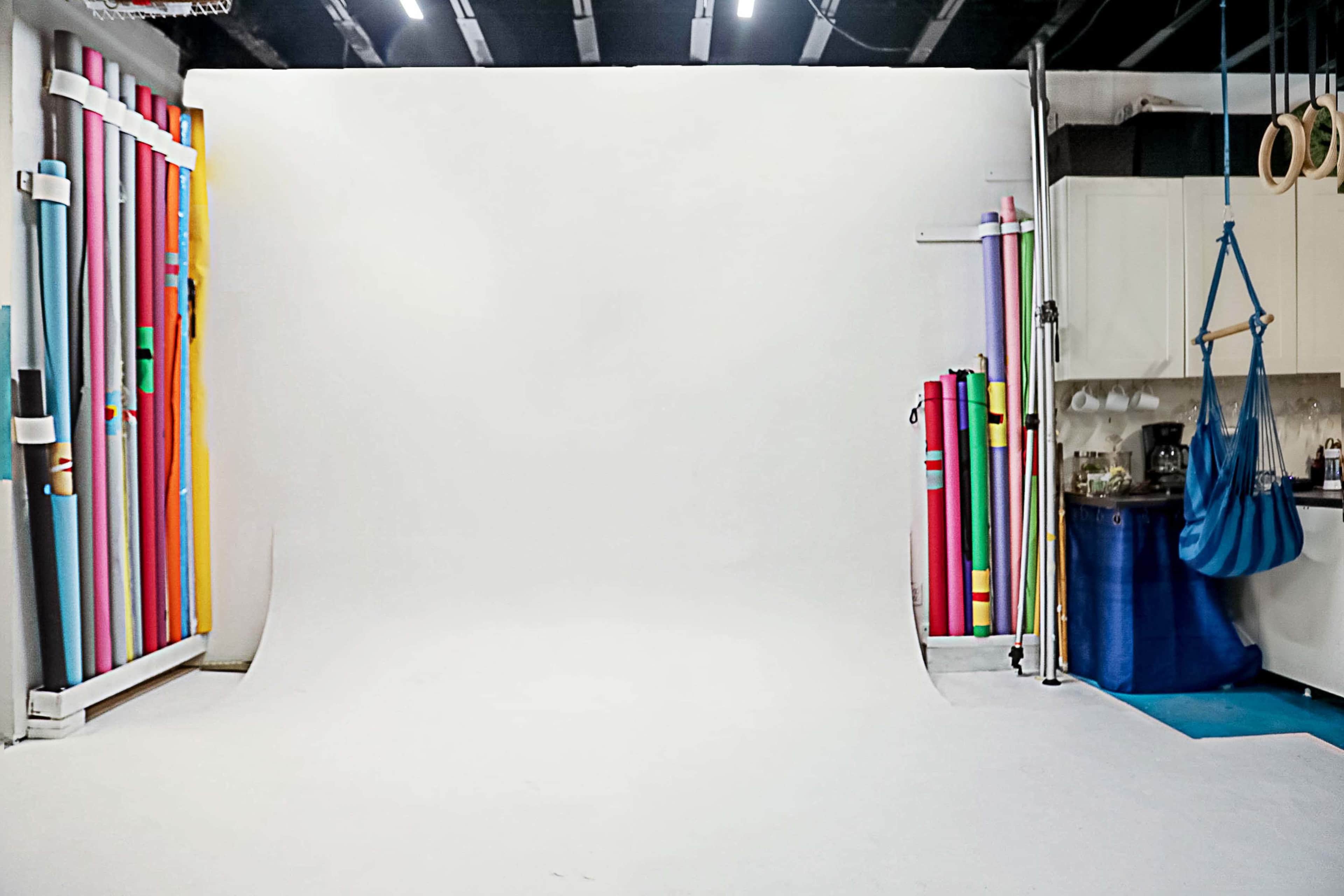The image shows a photography studio with a white backdrop and various colorful rolled backdrops stored along one side.
