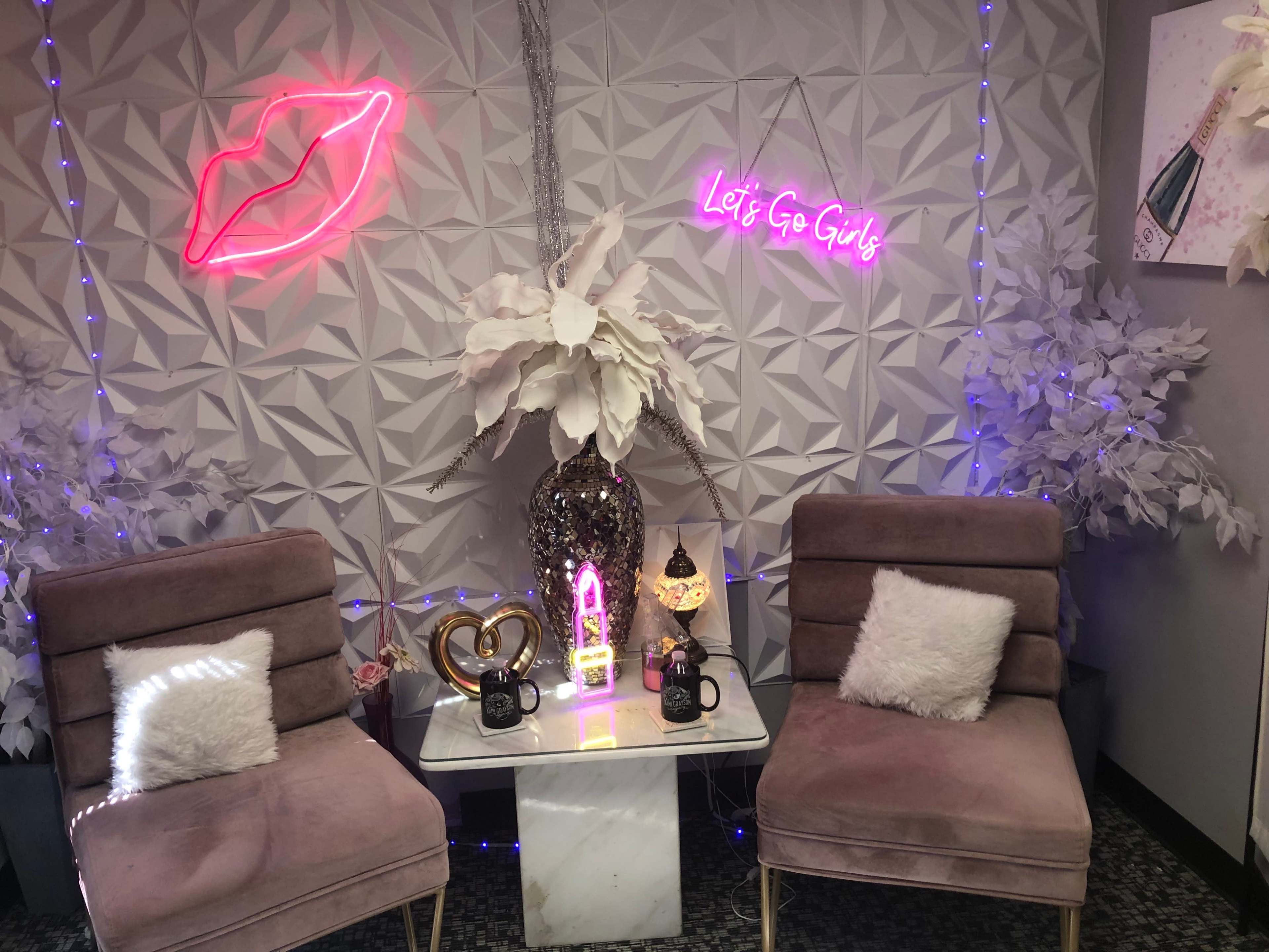 The image features a stylish seating area with two pink chairs, a white marble table, and neon signs reading "Let's Go Girls" and a pair of lips, surrounded by decorative flowers and soft lighting.