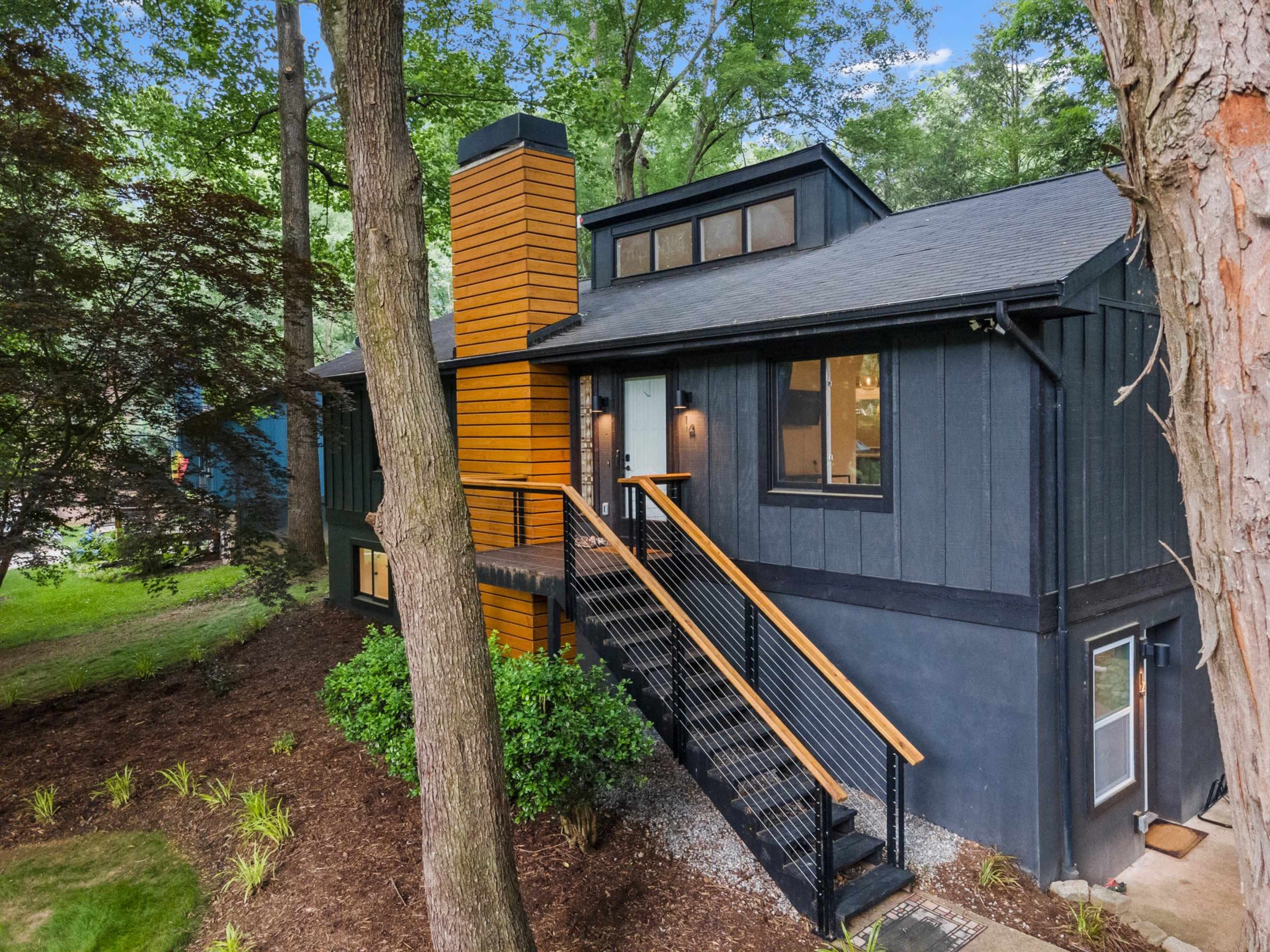 A modern black house with orange accents features a wooden staircase and a chimney, set among lush greenery.