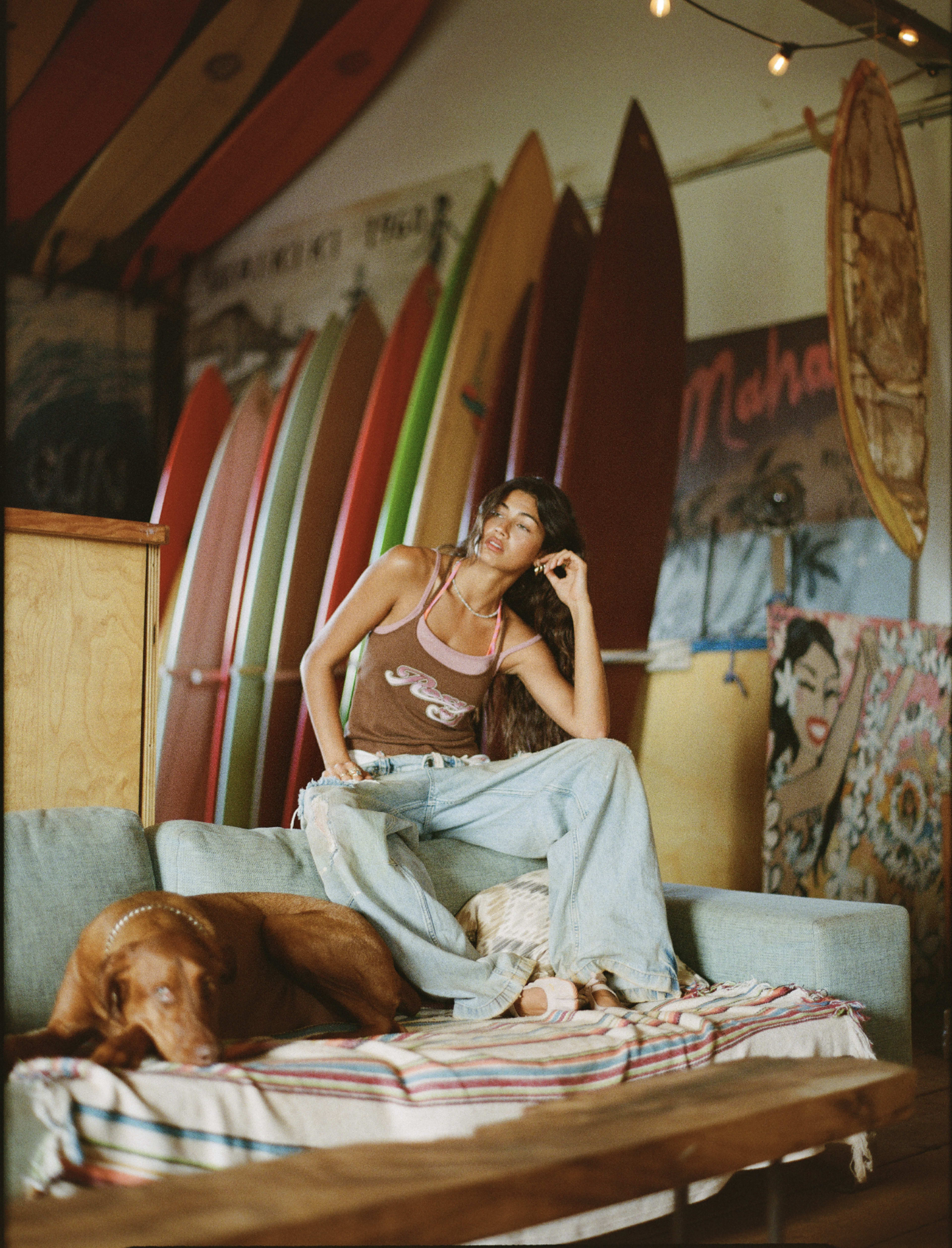 A woman sits on a couch with a dog, surrounded by colorful surfboards and artwork in a coastal-themed interior.