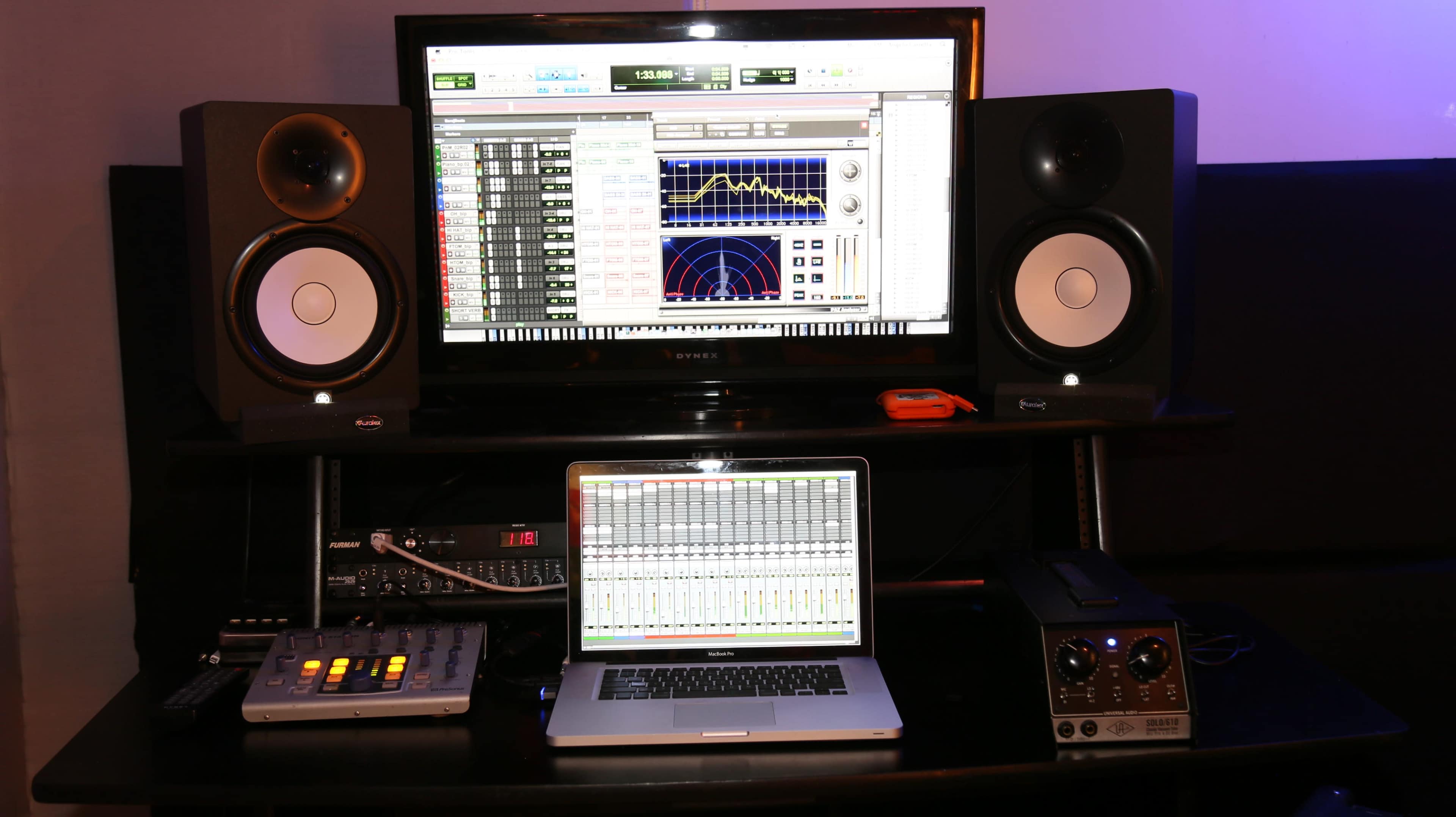 A music production setup features a computer monitor displaying audio software, two studio monitors, and a laptop with mixing controls on a desk.