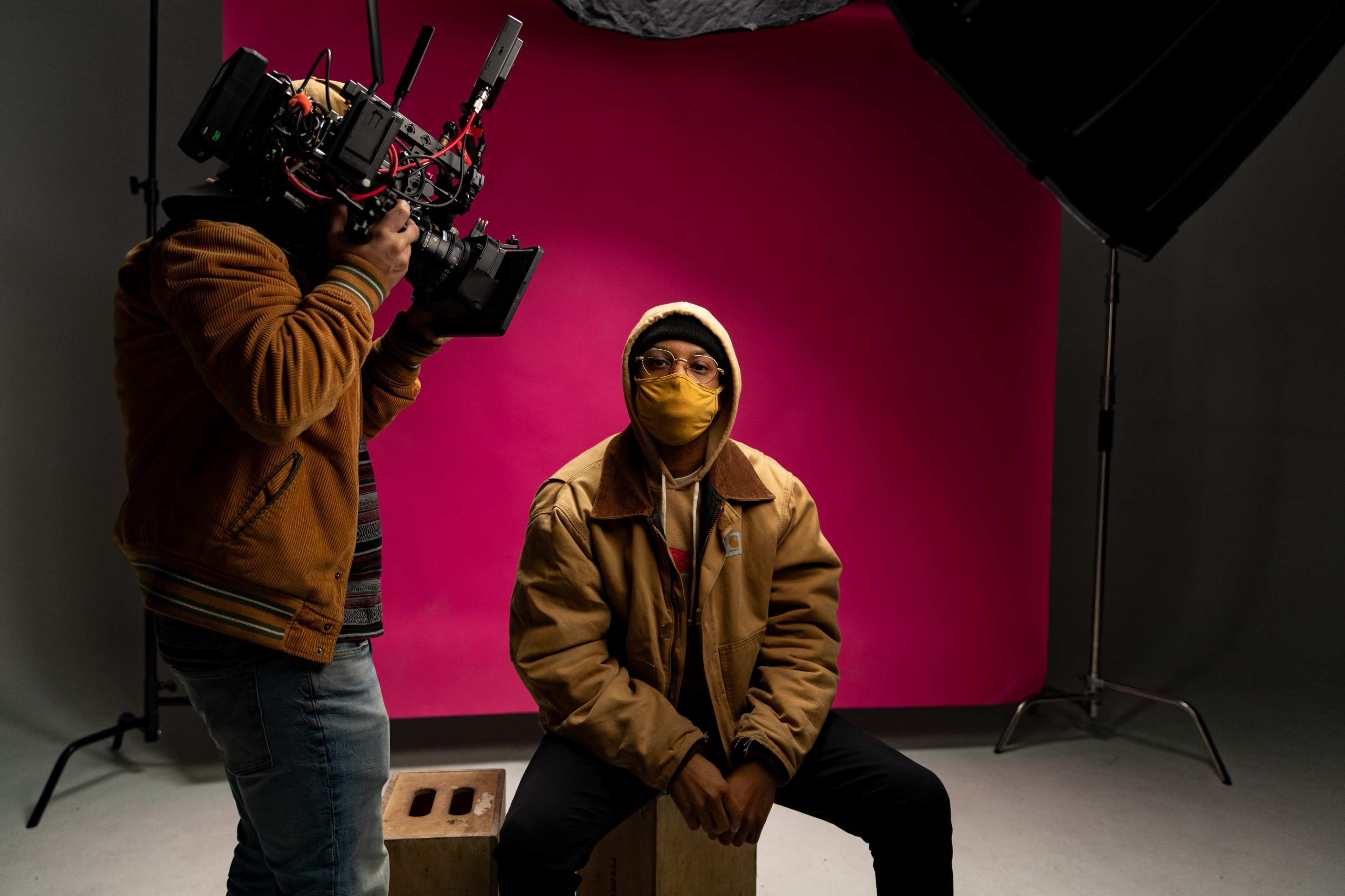 A photographer is capturing an image of a man wearing a yellow mask and a brown jacket, seated on a wooden block in front of a pink backdrop.