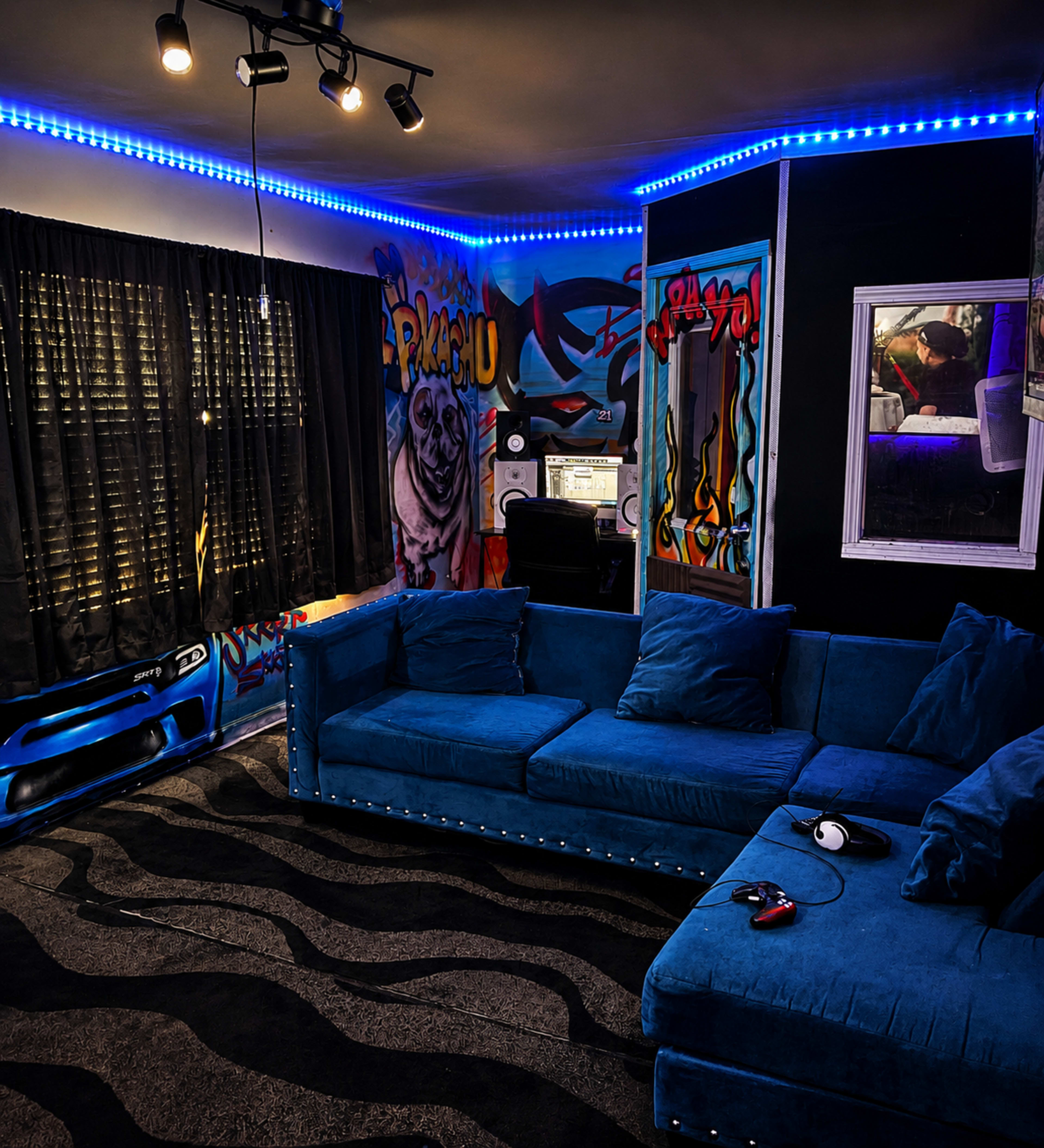The image shows a vibrant room featuring a blue sectional couch, graffiti-covered walls, a desk with a computer, and a large window covered by black curtains.