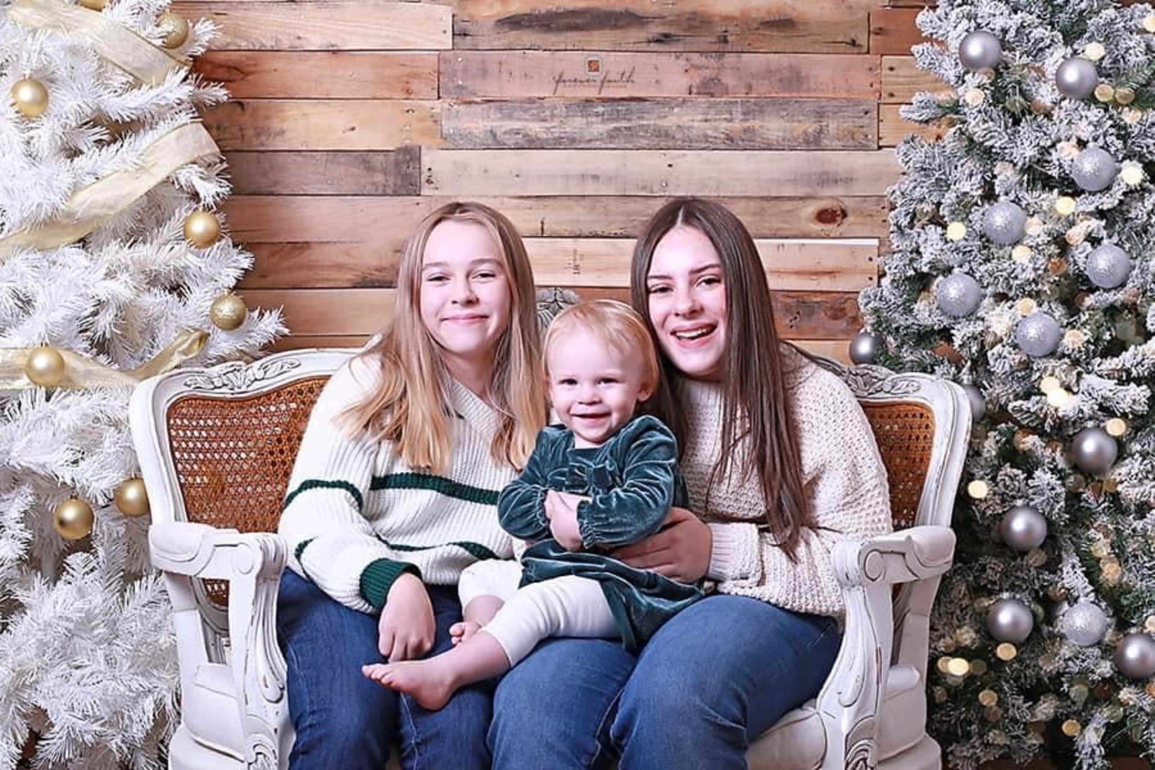 Two girls sit on a sofa beside a toddler, with decorated Christmas trees in the background.