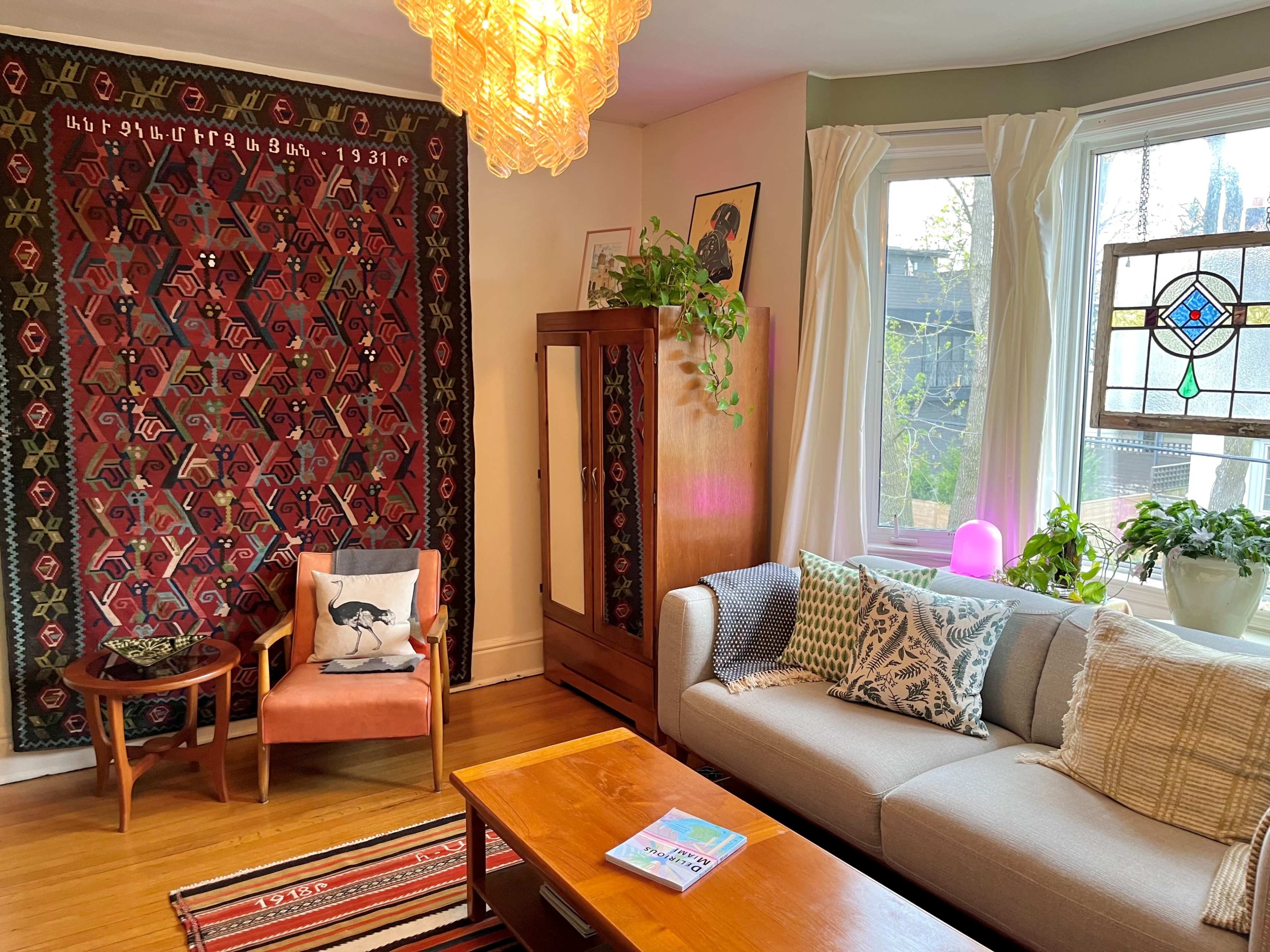 The scene depicts a cozy living room with a patterned wall tapestry, a light fixture hanging from the ceiling, a sofa adorned with decorative pillows, and a mix of plants near a large window.