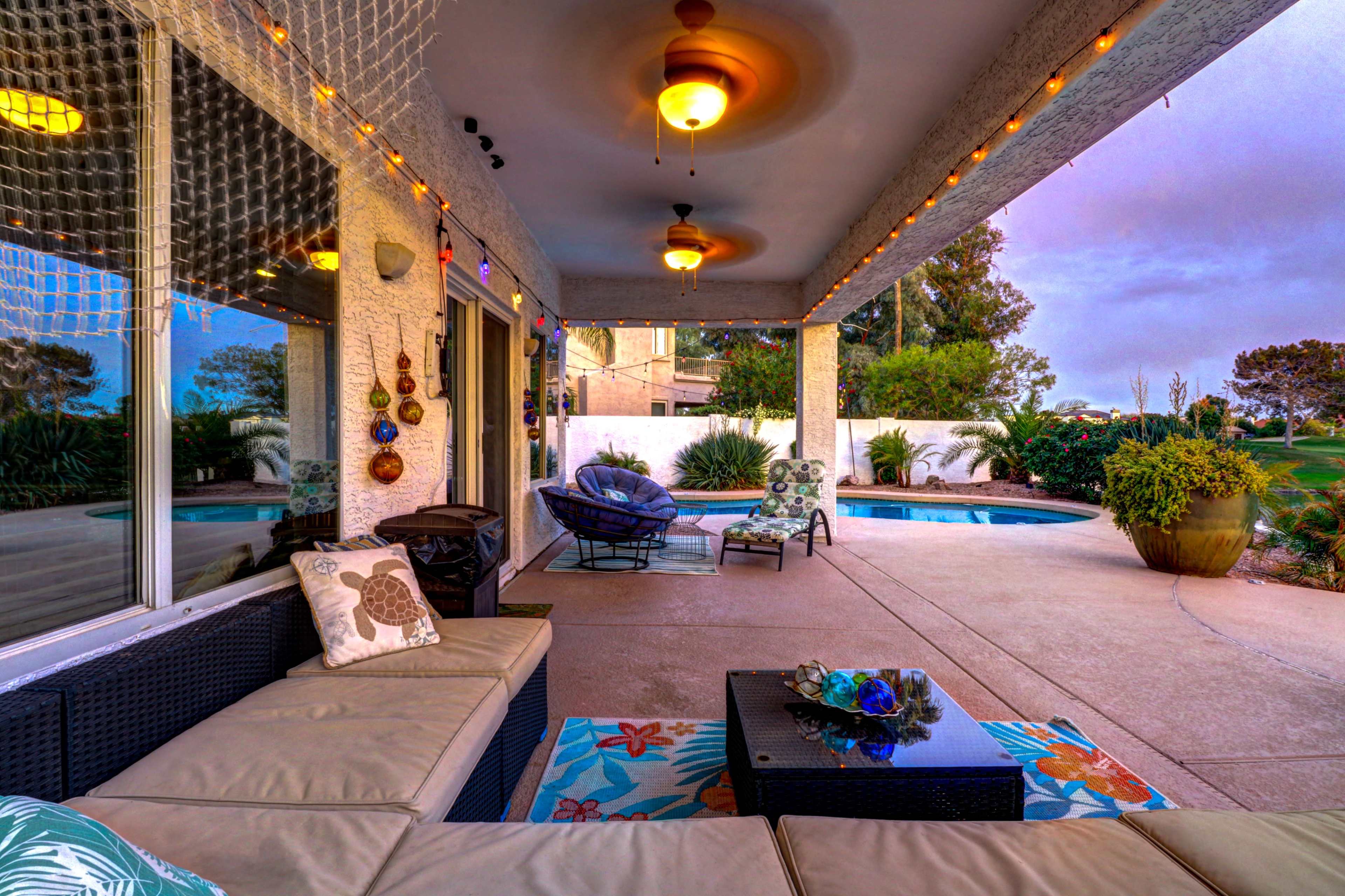 The image shows a patio area with a sectional sofa, decorative lights, and a view of a pool surrounded by greenery.