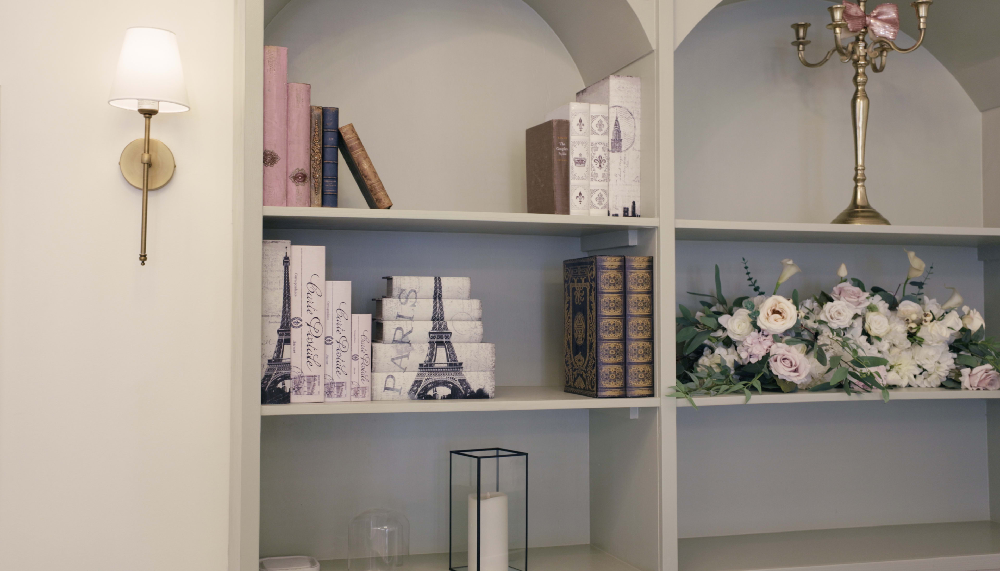 The image shows a bookshelf with various decorative items, including stacked books, a decorative box, a lantern, and a floral arrangement.