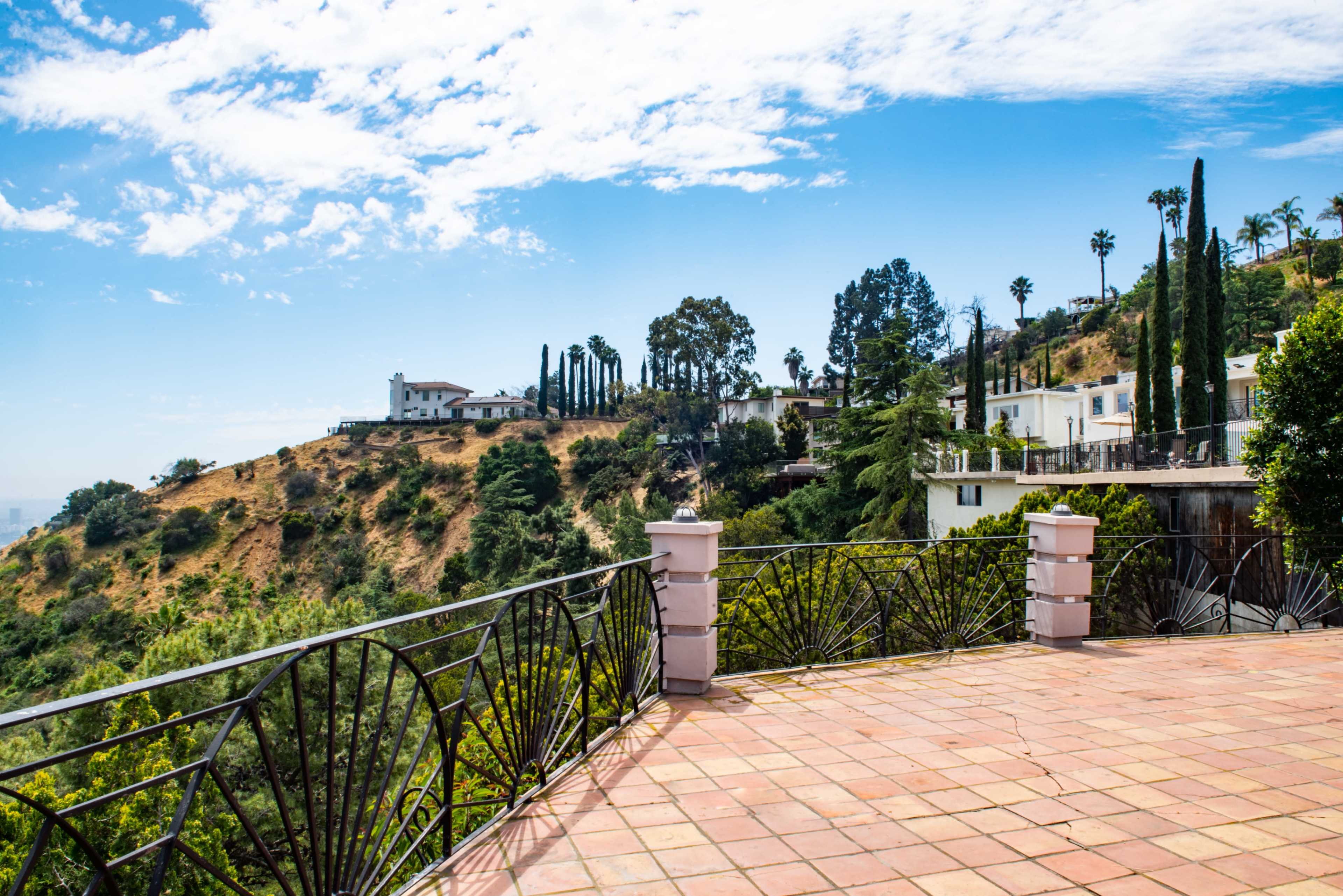 The image shows a scenic view from a terrace overlooking a hillside dotted with trees and large houses under a blue sky with scattered clouds.