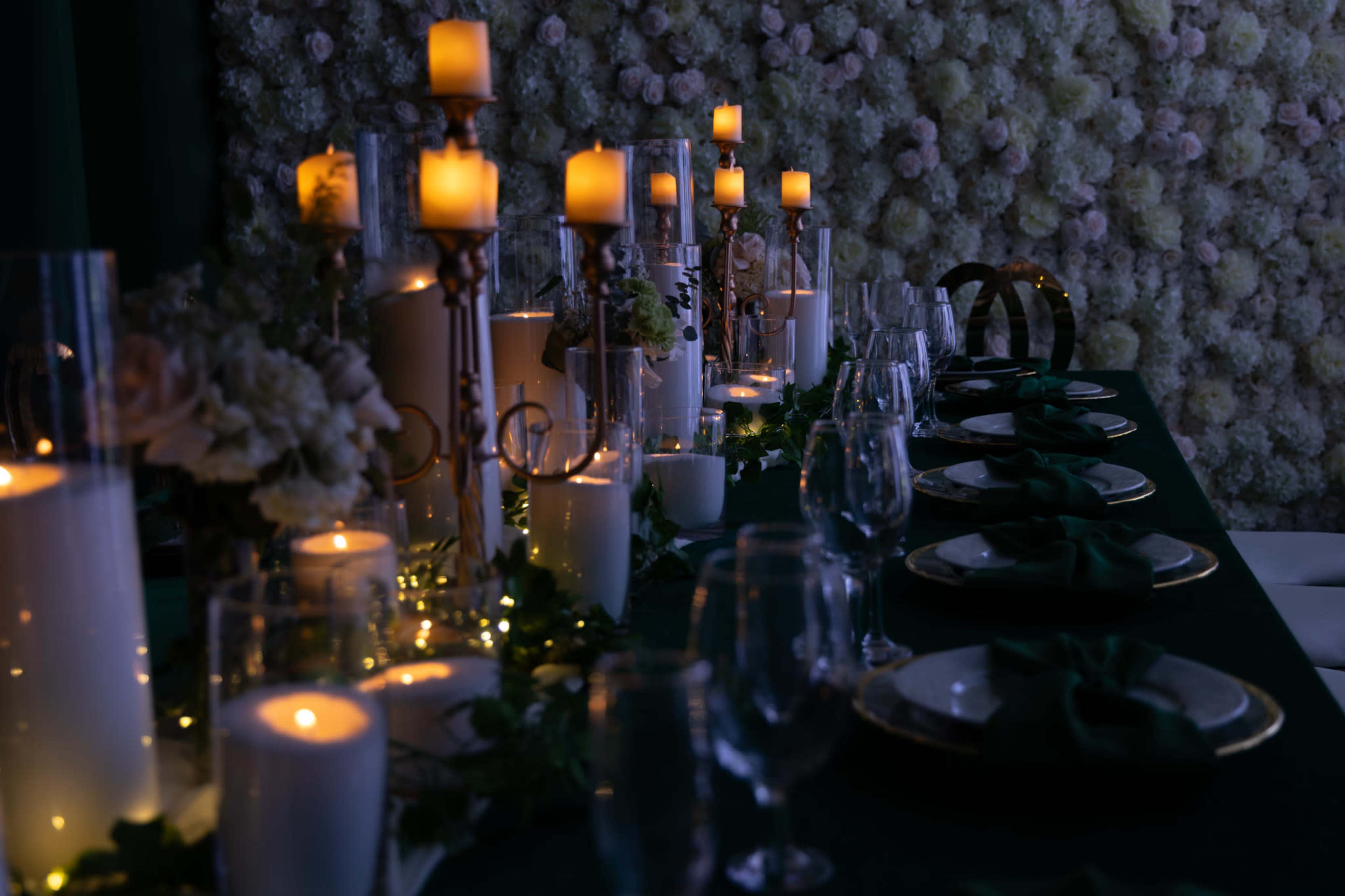 A dimly lit table is elegantly set with candles, glassware, and dark green napkins, against a backdrop of a floral wall.