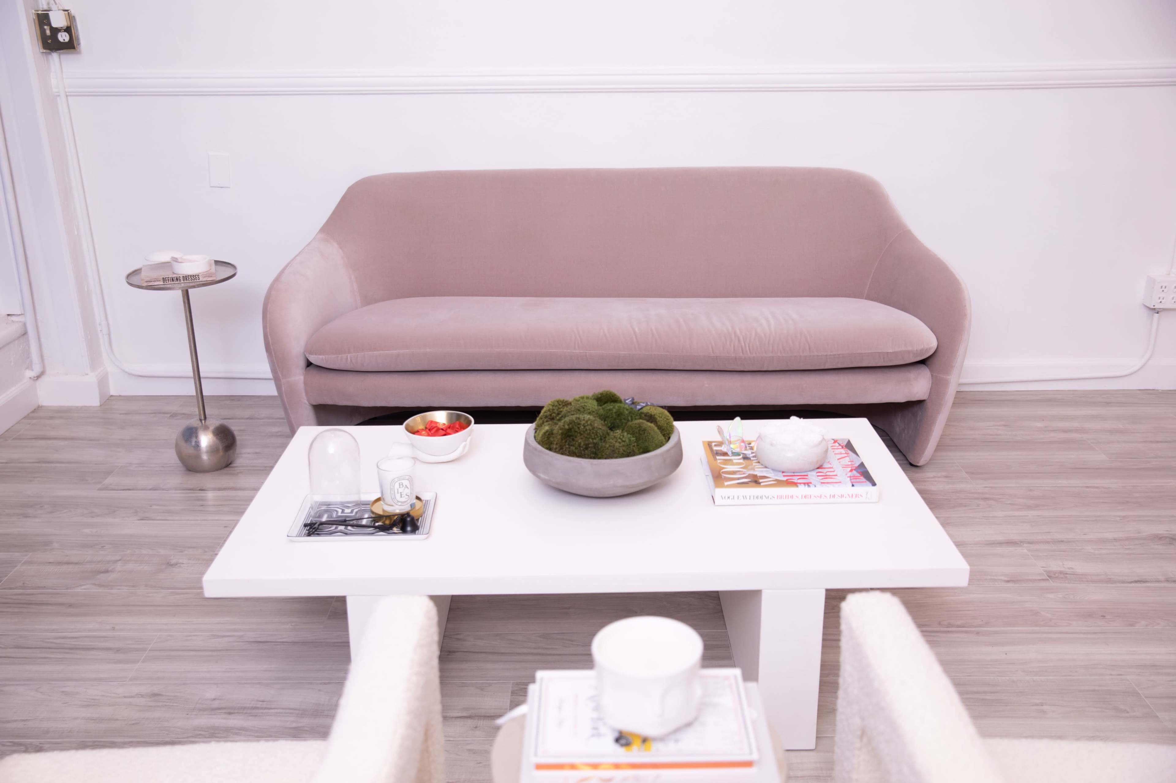 A soft pink sofa positioned facing a white coffee table, which holds a bowl of green moss, a small dish of fruit, and a stack of magazines, all set against a light-colored wooden floor.