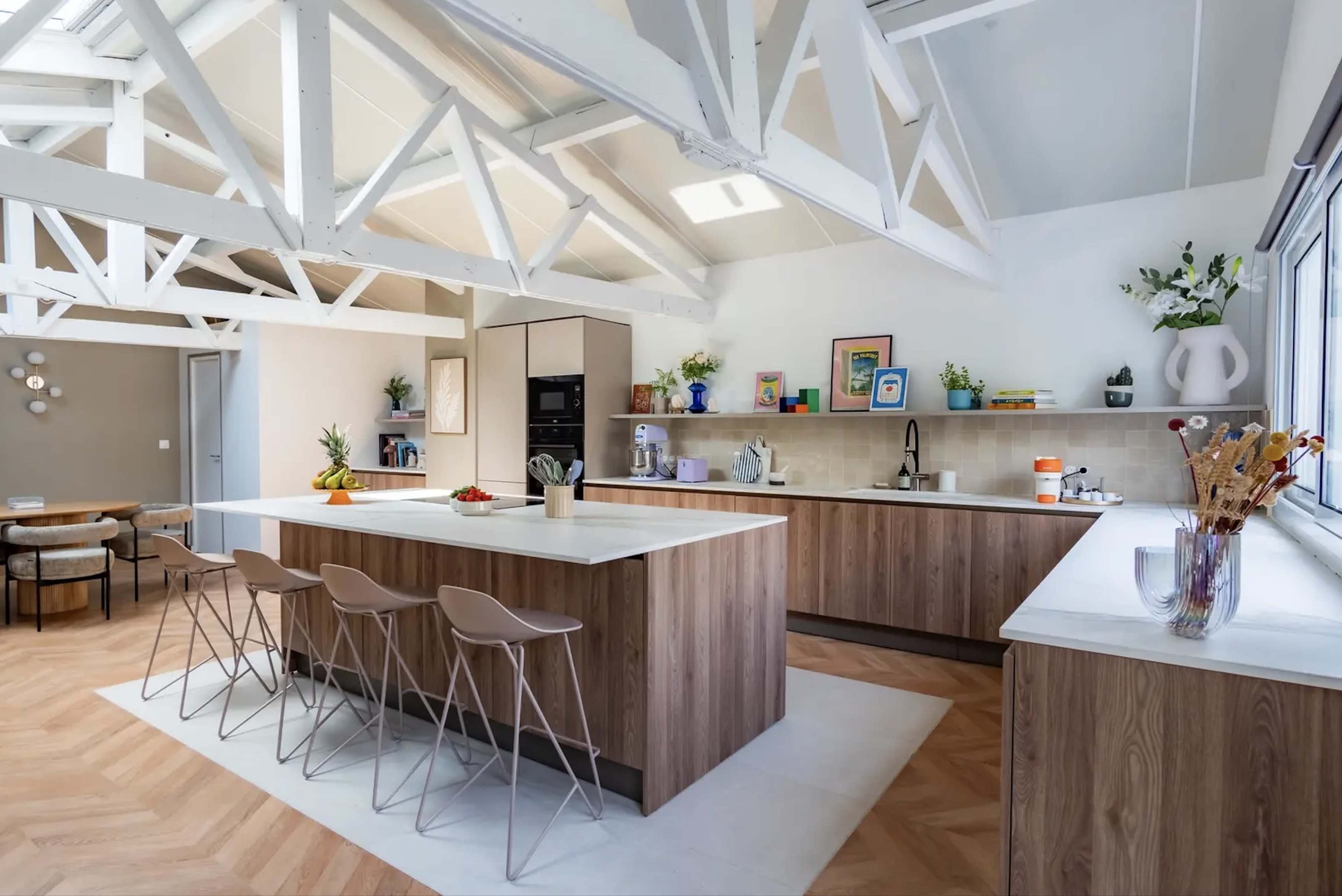 The image shows a modern kitchen with a large island, wooden cabinetry, and high ceilings supported by white beams.