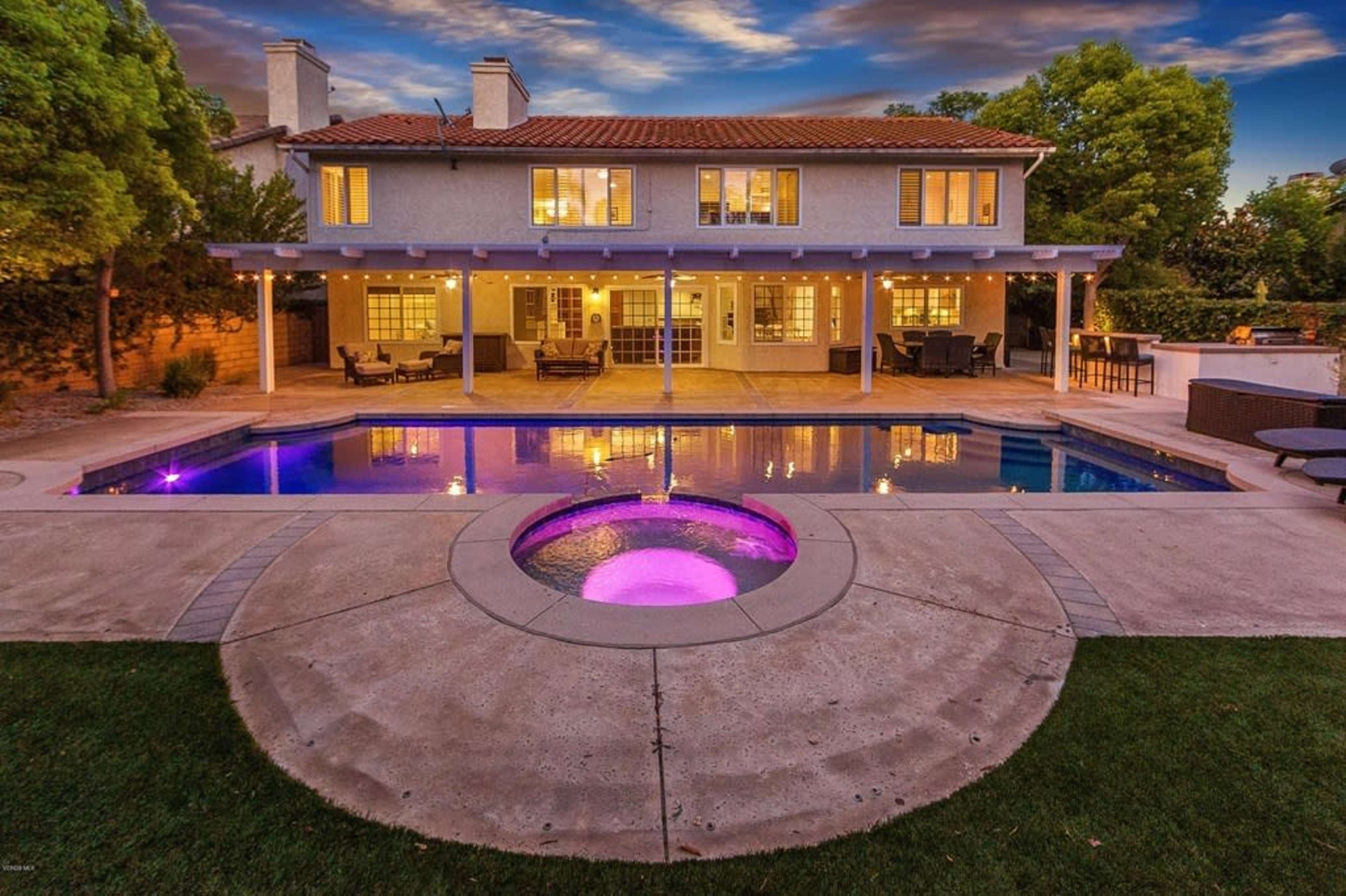 A two-story house with a tiled roof overlooks a backyard featuring a swimming pool and a hot tub surrounded by landscaped greenery.