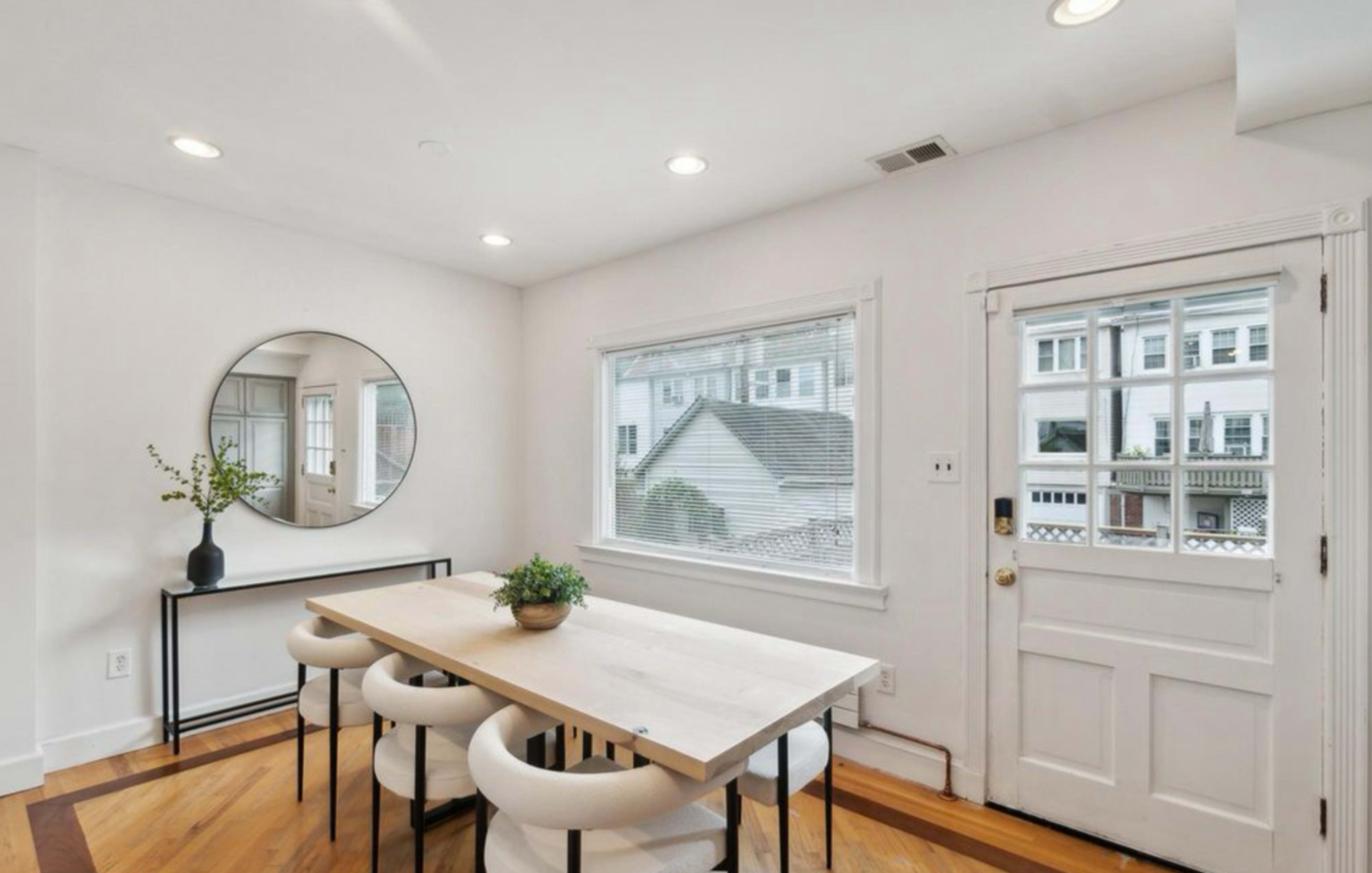A dining area features a light wood table surrounded by four white upholstered chairs, with a large round mirror and a window providing natural light.