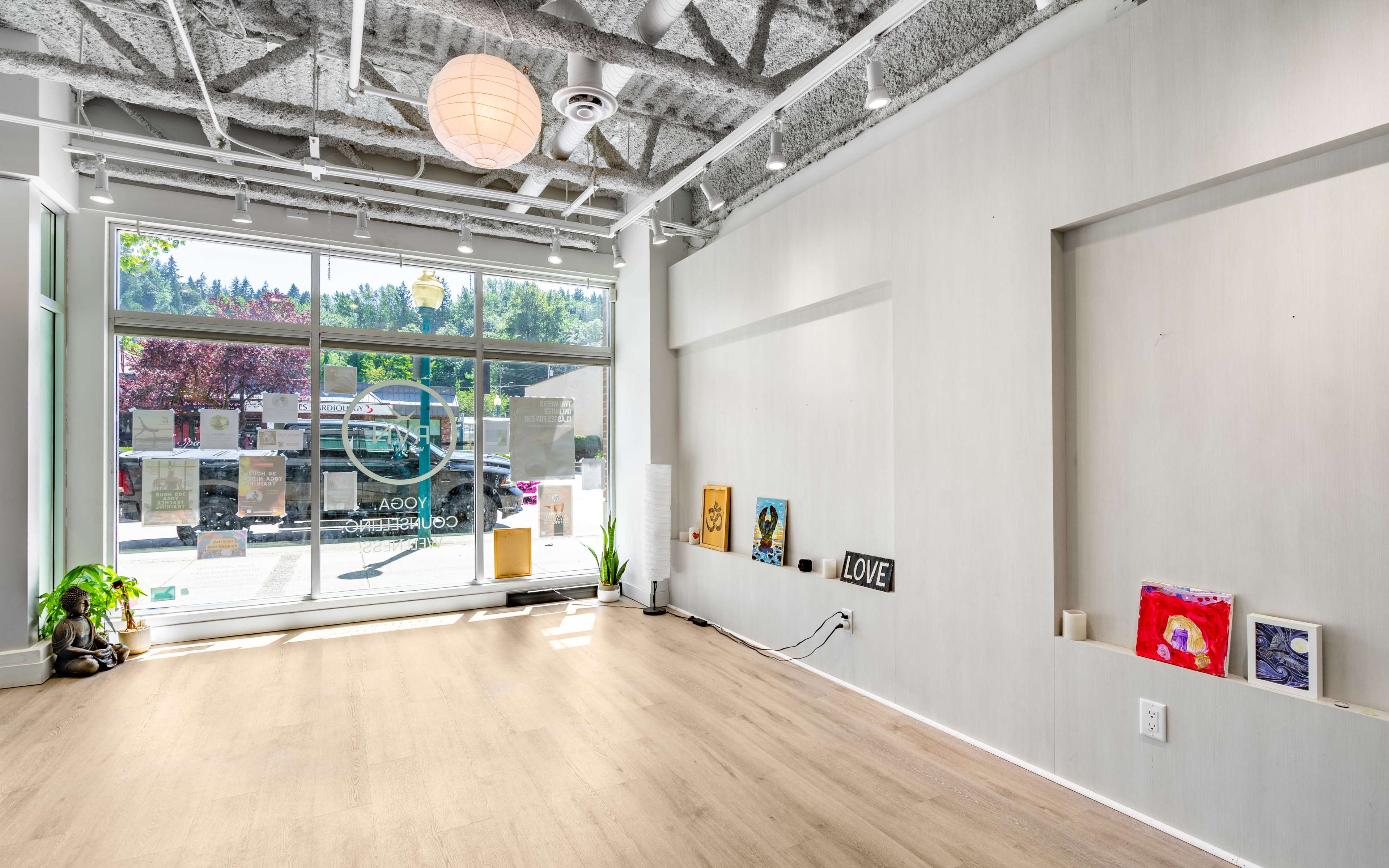 A bright, minimalist interior of a gallery or studio with large windows facing the street, wooden flooring, and a few art pieces displayed on the walls and shelves.