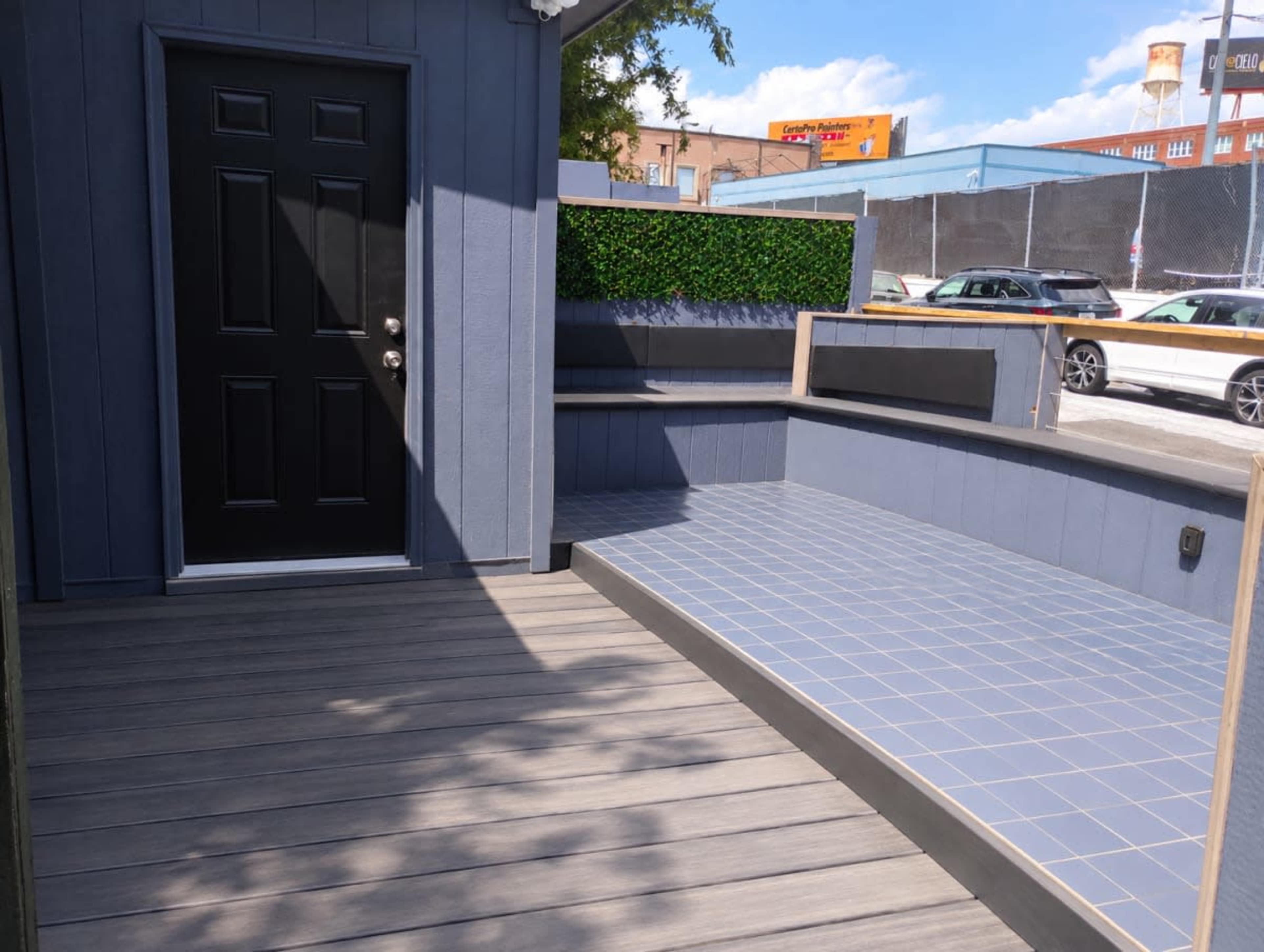 The image shows a modern outdoor space featuring a wooden deck, a gray wall with a black door, and a tiled seating area with a hedge backdrop.