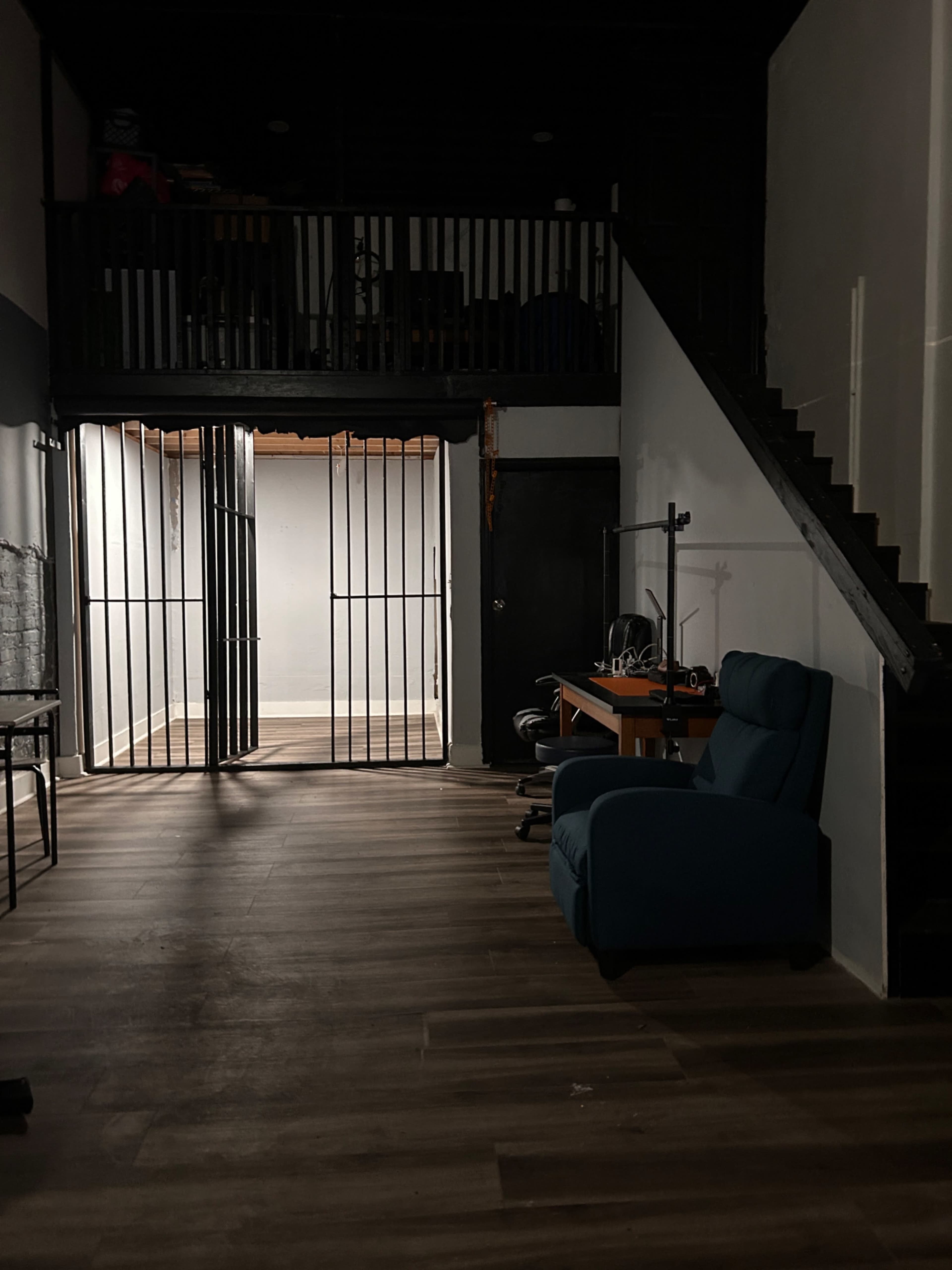The image shows a dimly lit room with a blue armchair, a desk with office equipment, and a staircase leading to a wooden loft, all separated by metal bars on the doorway.