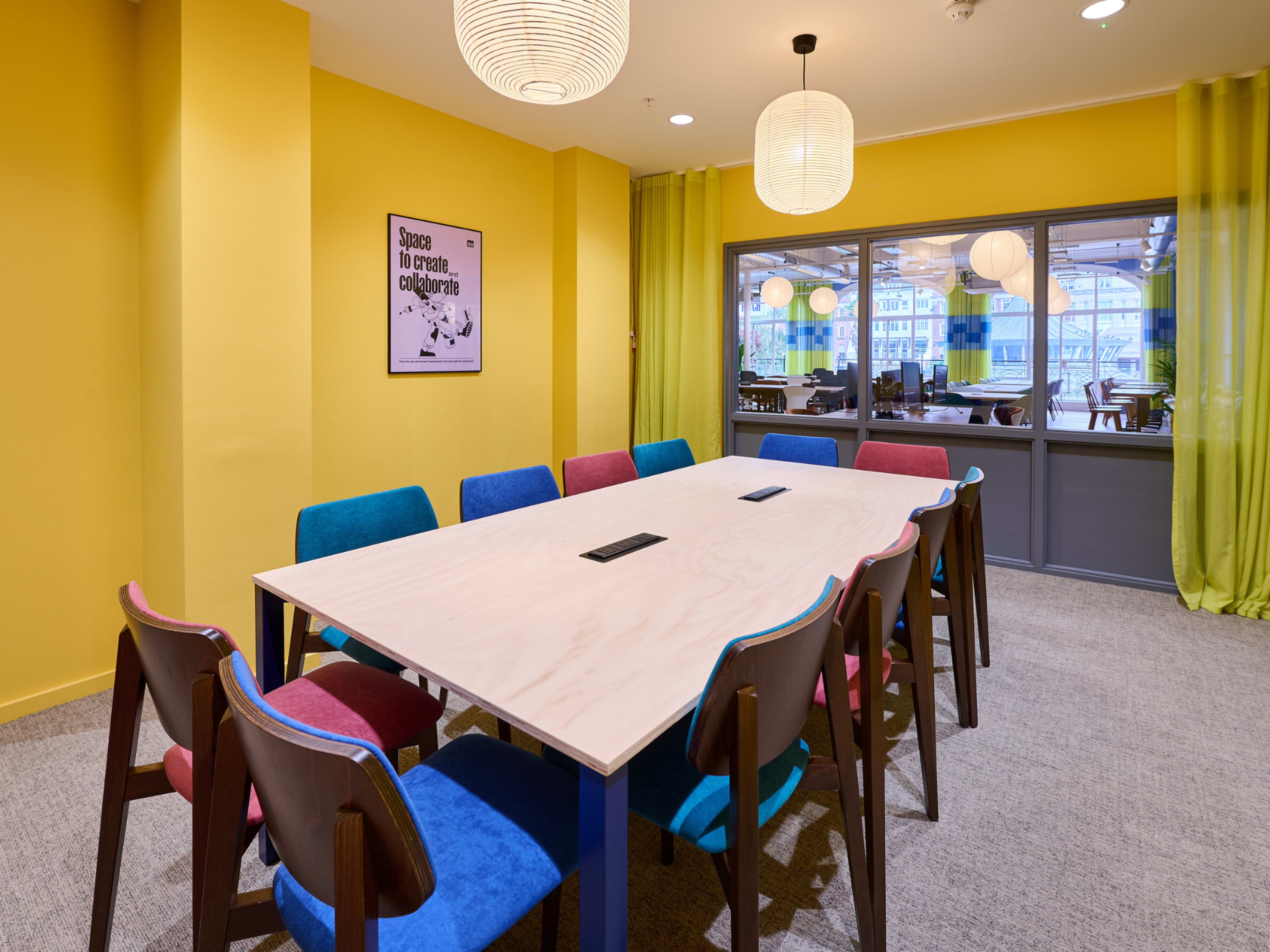 A conference room features a large wooden table surrounded by colorful chairs, with yellow walls and large windows letting in natural light.