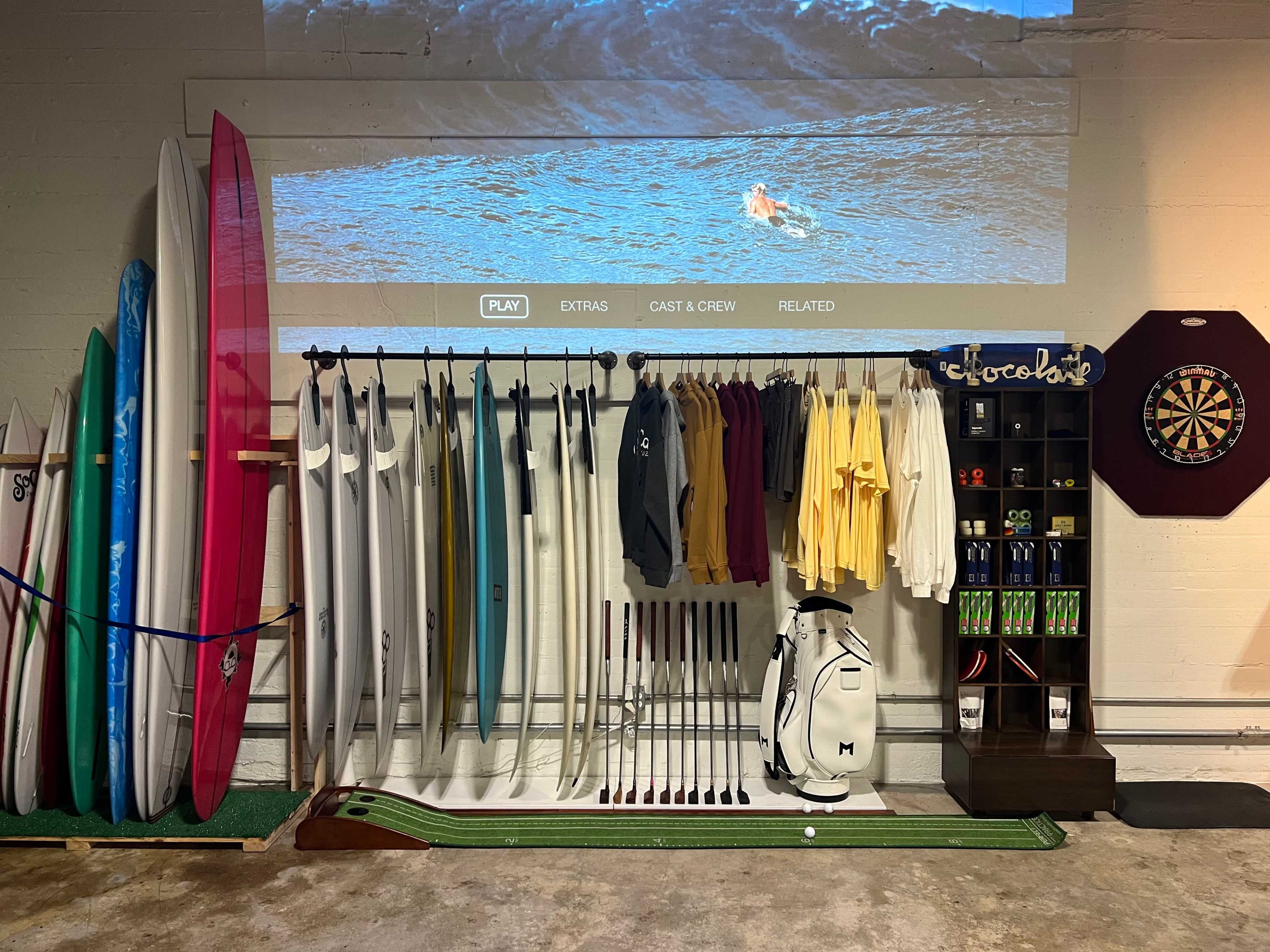 The image shows a collection of surfboards and golf equipment arranged in a garage-like setting, with a screen displaying a video in the background and a dartboard on the side.