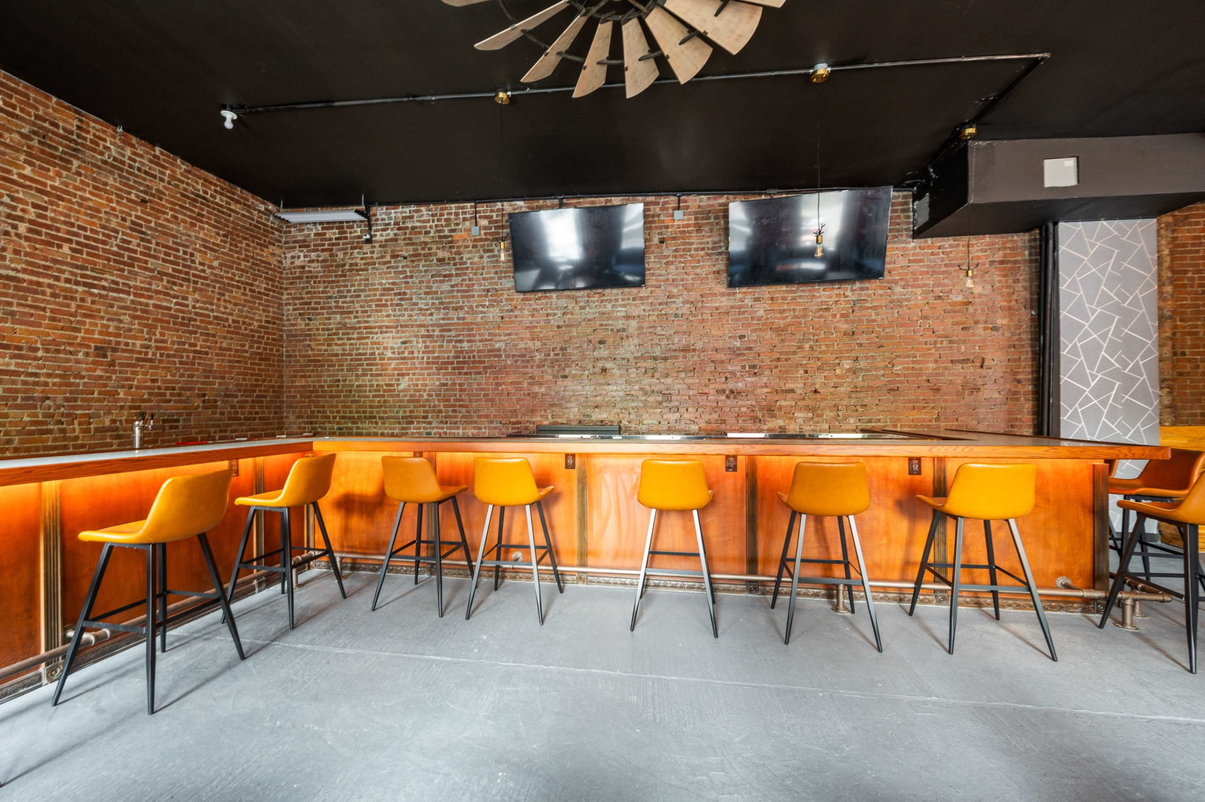 A modern bar area with a wooden counter, orange bar stools, exposed brick walls, and two flat-screen televisions mounted above.