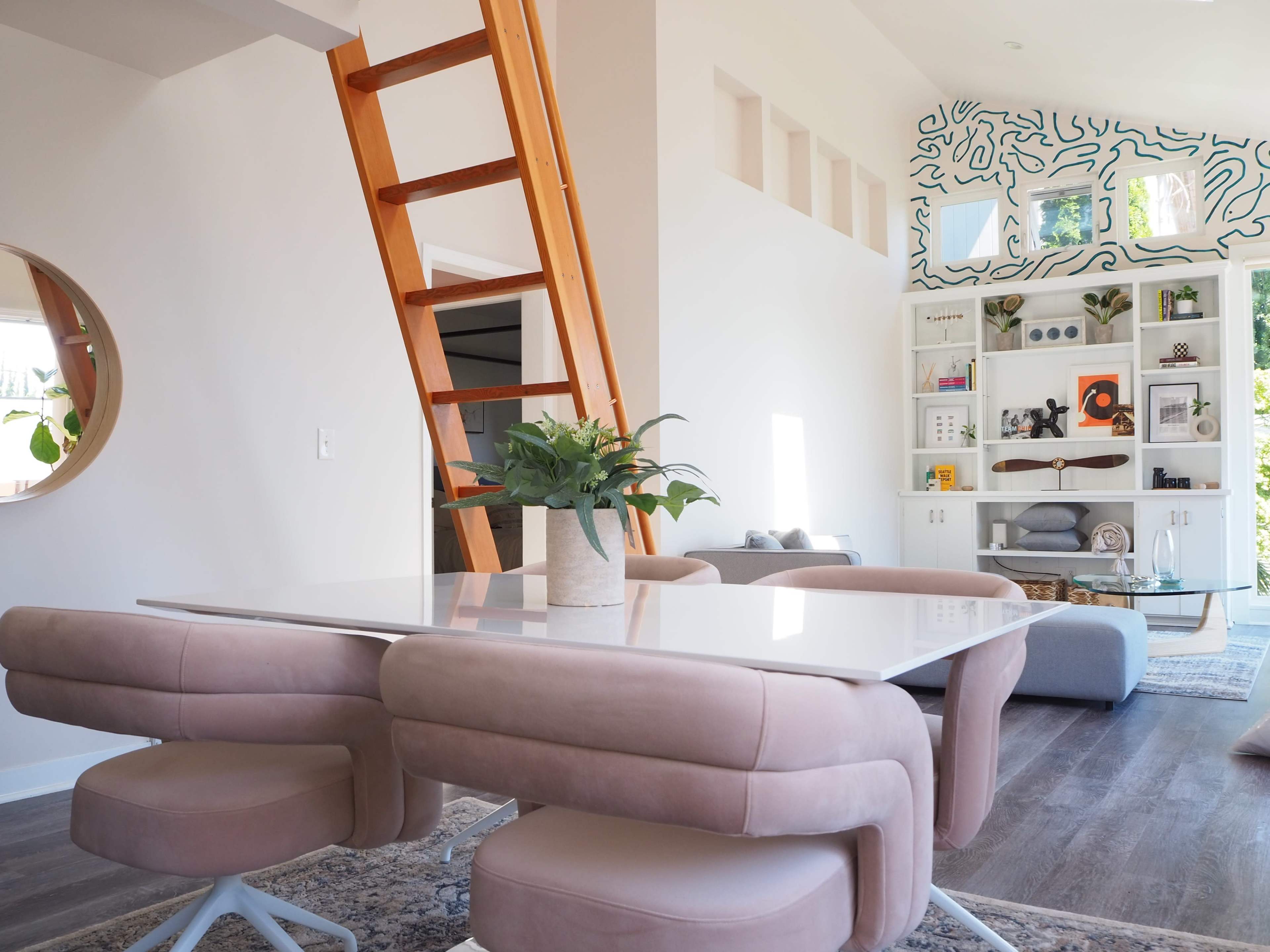 A modern living space features a dining table with pink chairs, a wooden ladder leading to an upper level, and a bookshelf against a wall with decorative accents.