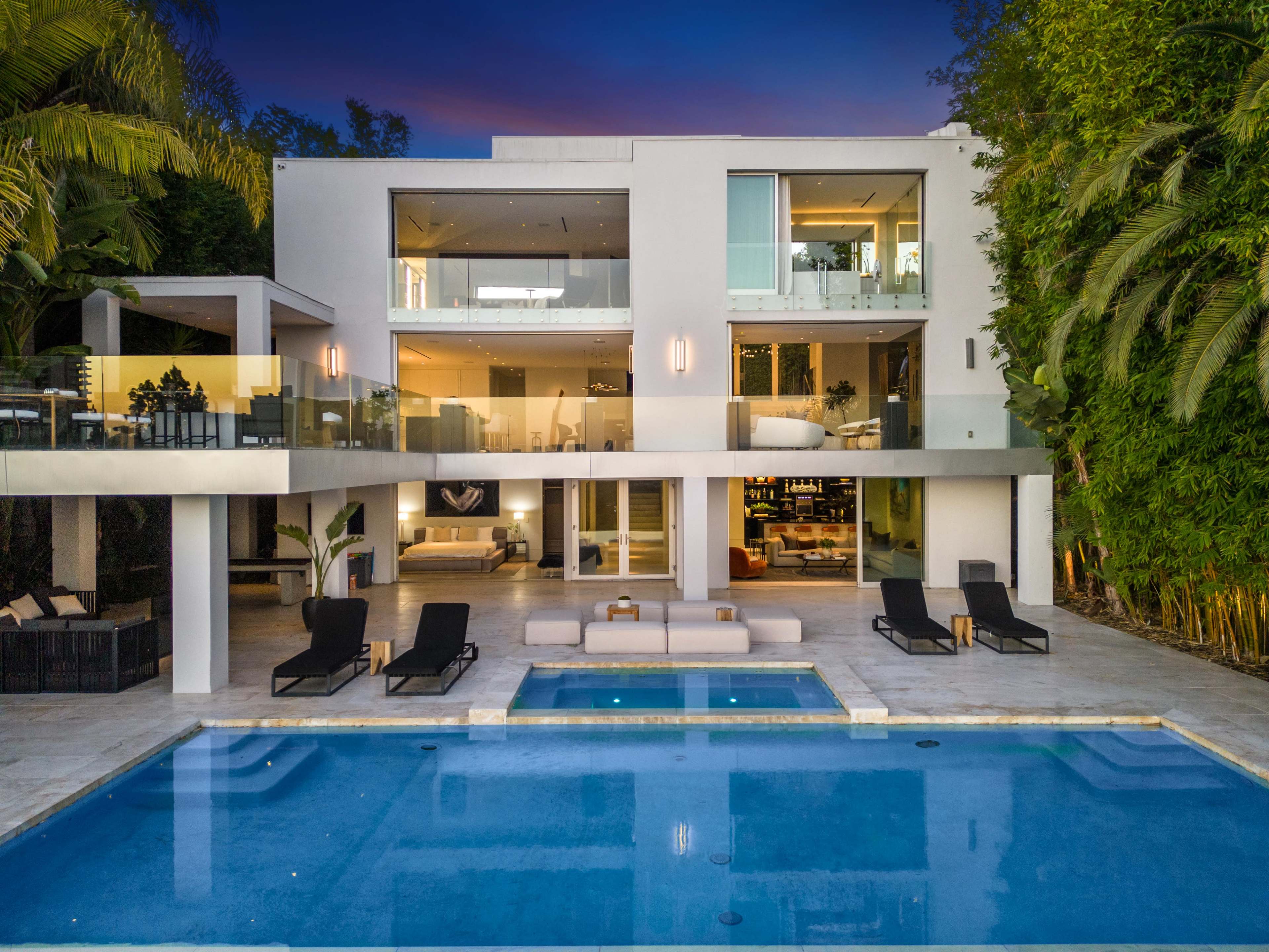 The image shows a modern two-story house with large windows, a swimming pool in the foreground, and landscaped greenery surrounding the property.