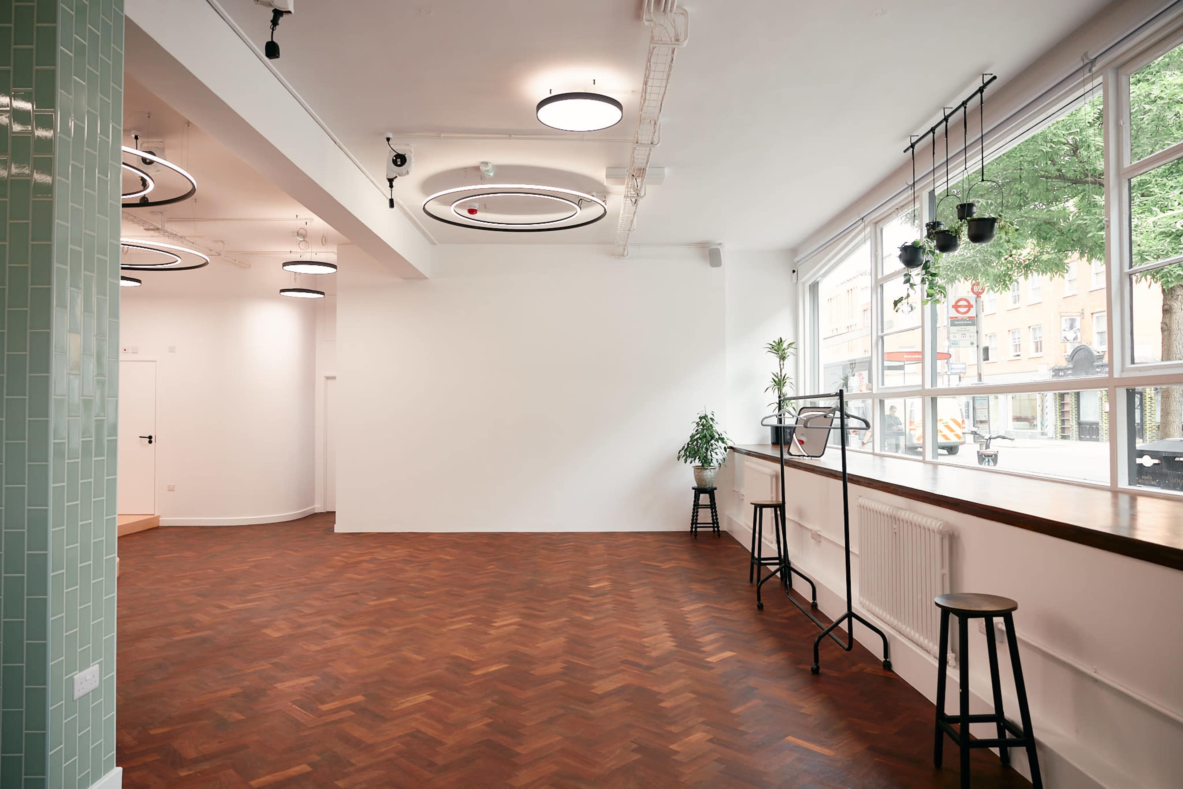An empty, modern space featuring wooden herringbone flooring, a wooden countertop with high stools, and large windows allowing natural light to enter.