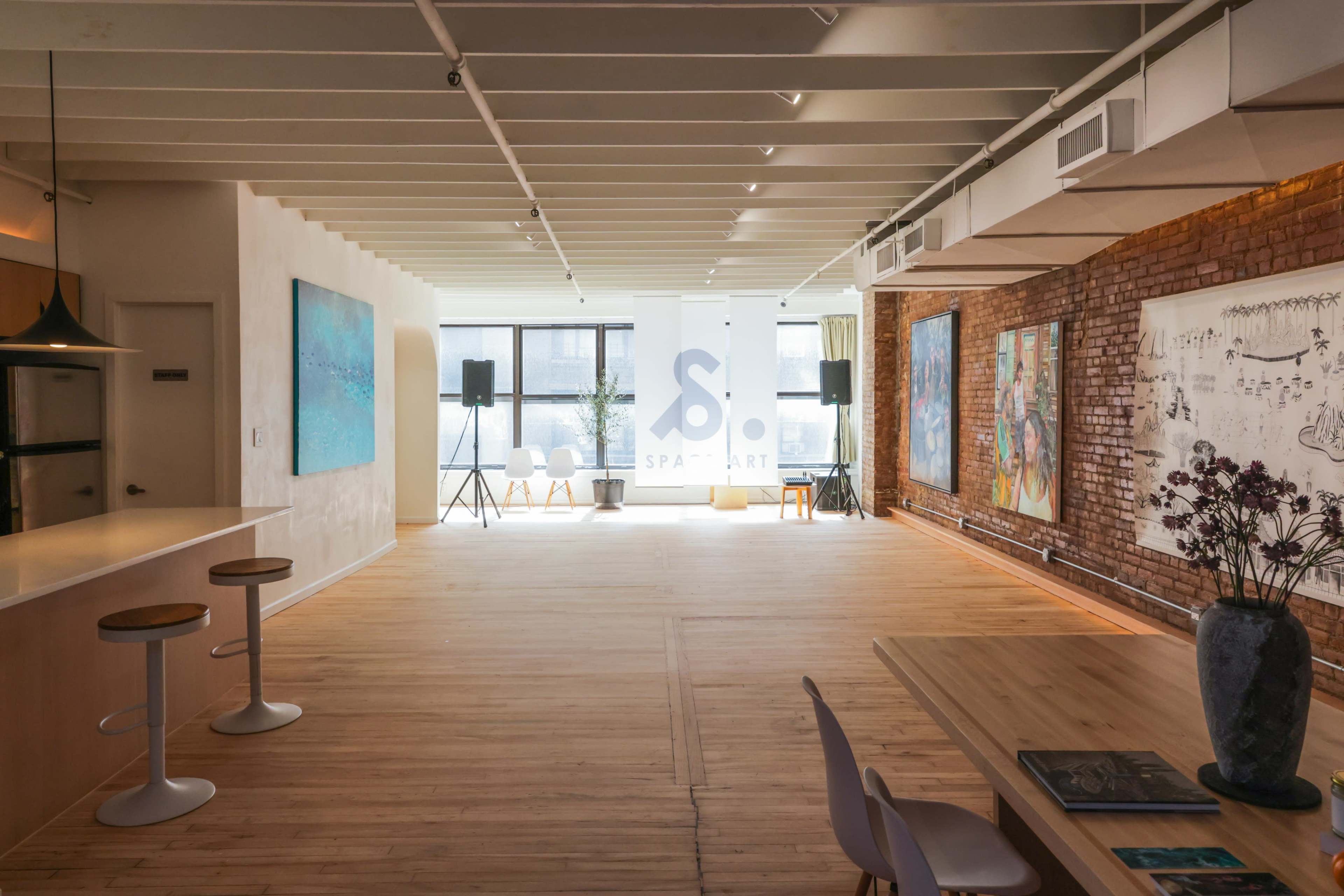 The interior of a spacious art studio features a wooden floor, large windows, and various artworks displayed on the walls, with seating and equipment for events.