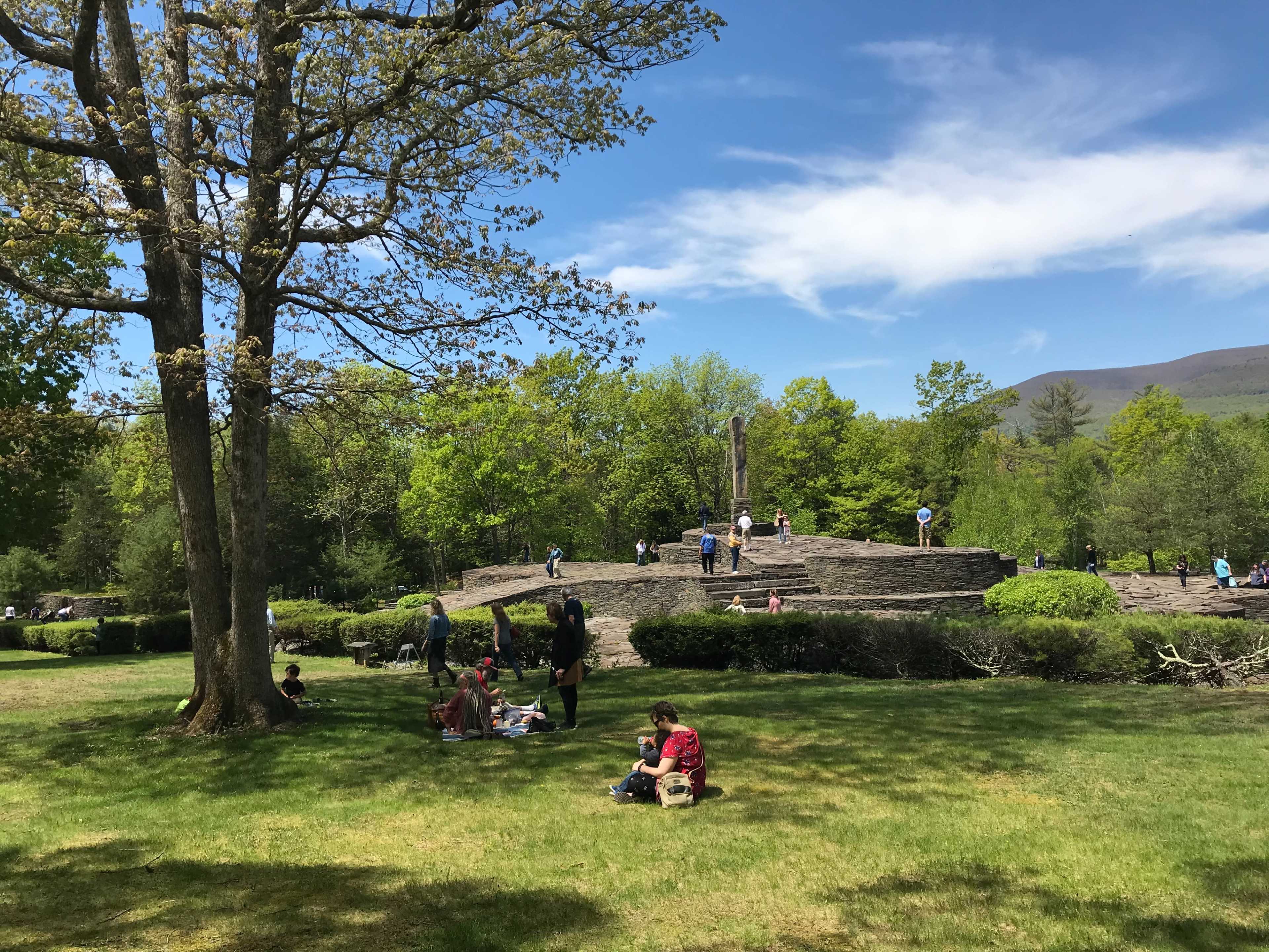 People are gathered on the grass in a park, with a large wooden structure and trees in the background under a partly cloudy sky.