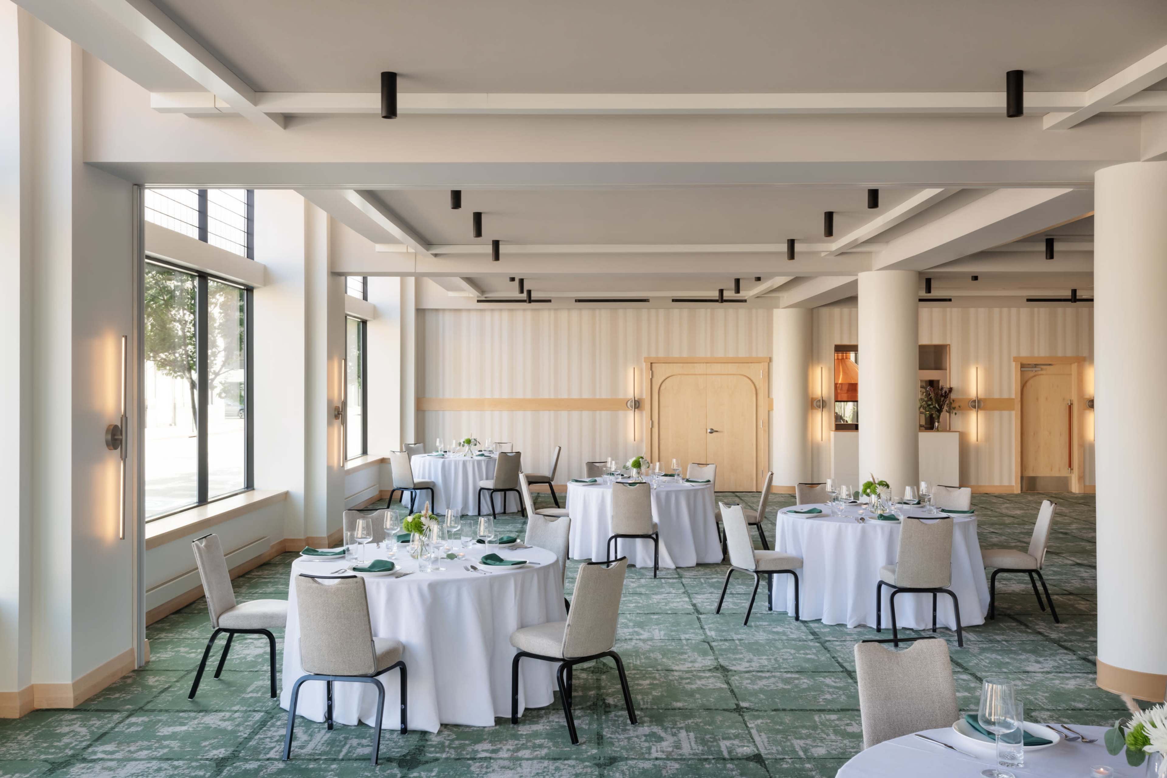 The image shows a modern dining room with multiple round tables set for a meal, surrounded by large windows that let in natural light.