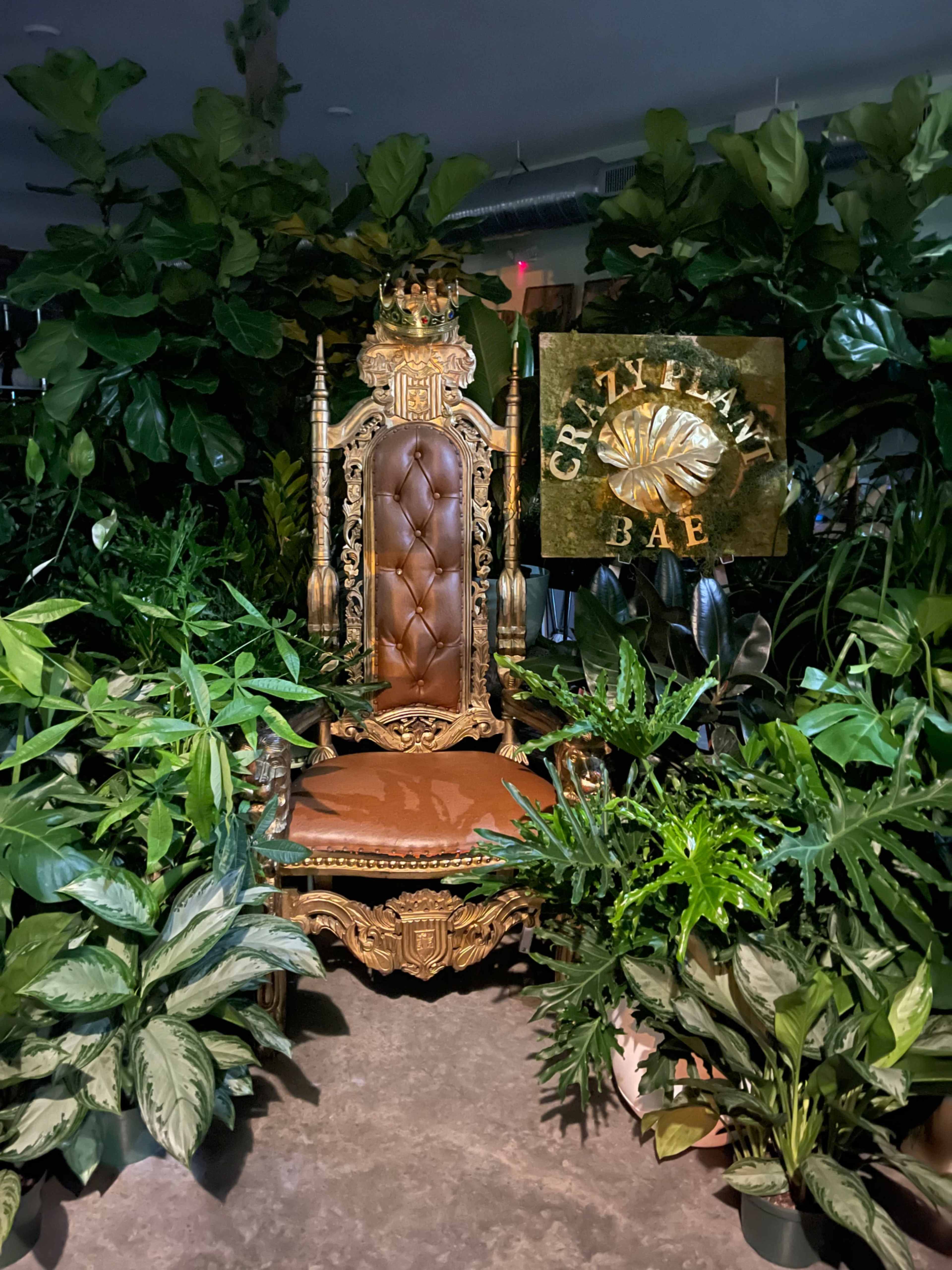 An ornate throne surrounded by lush greenery and a sign that reads "CRZY PLANT BAE."