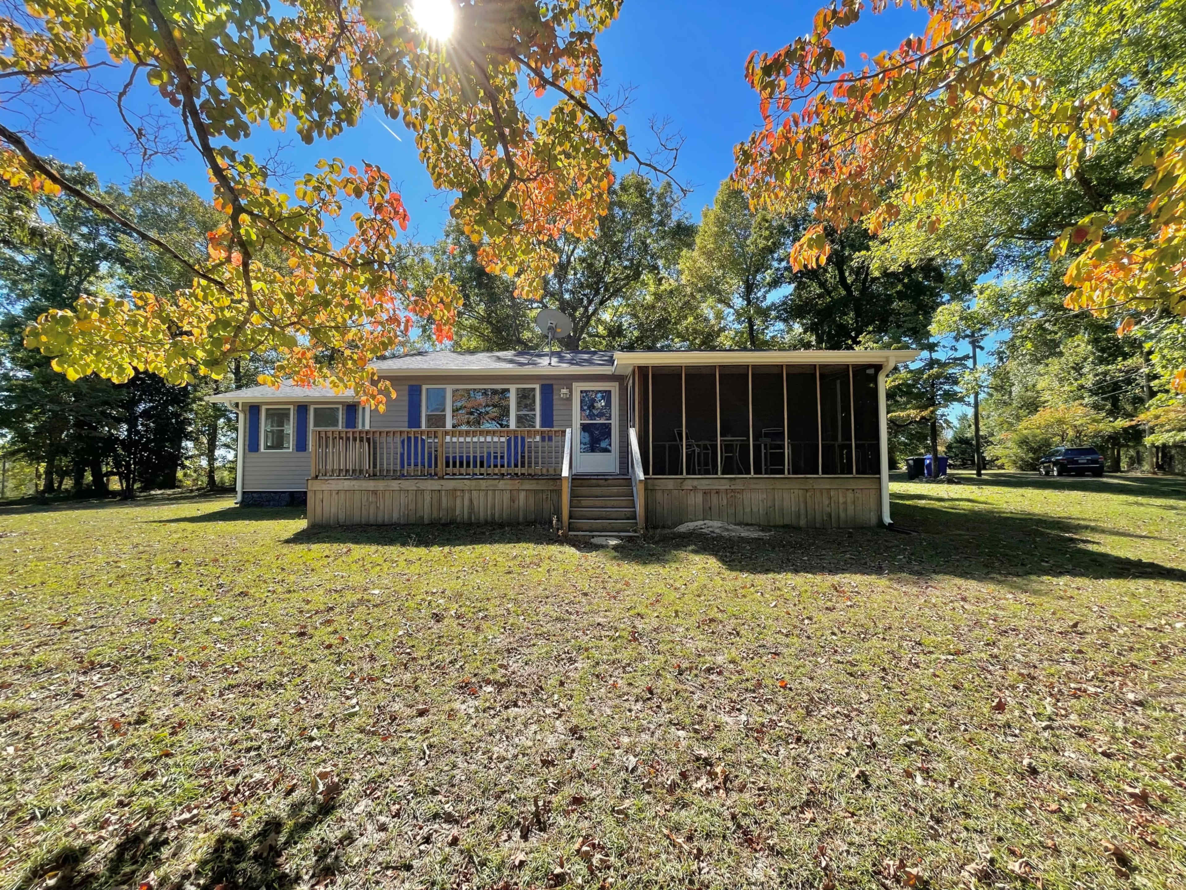A single-story house with a screened porch and a front deck is surrounded by grass and trees.