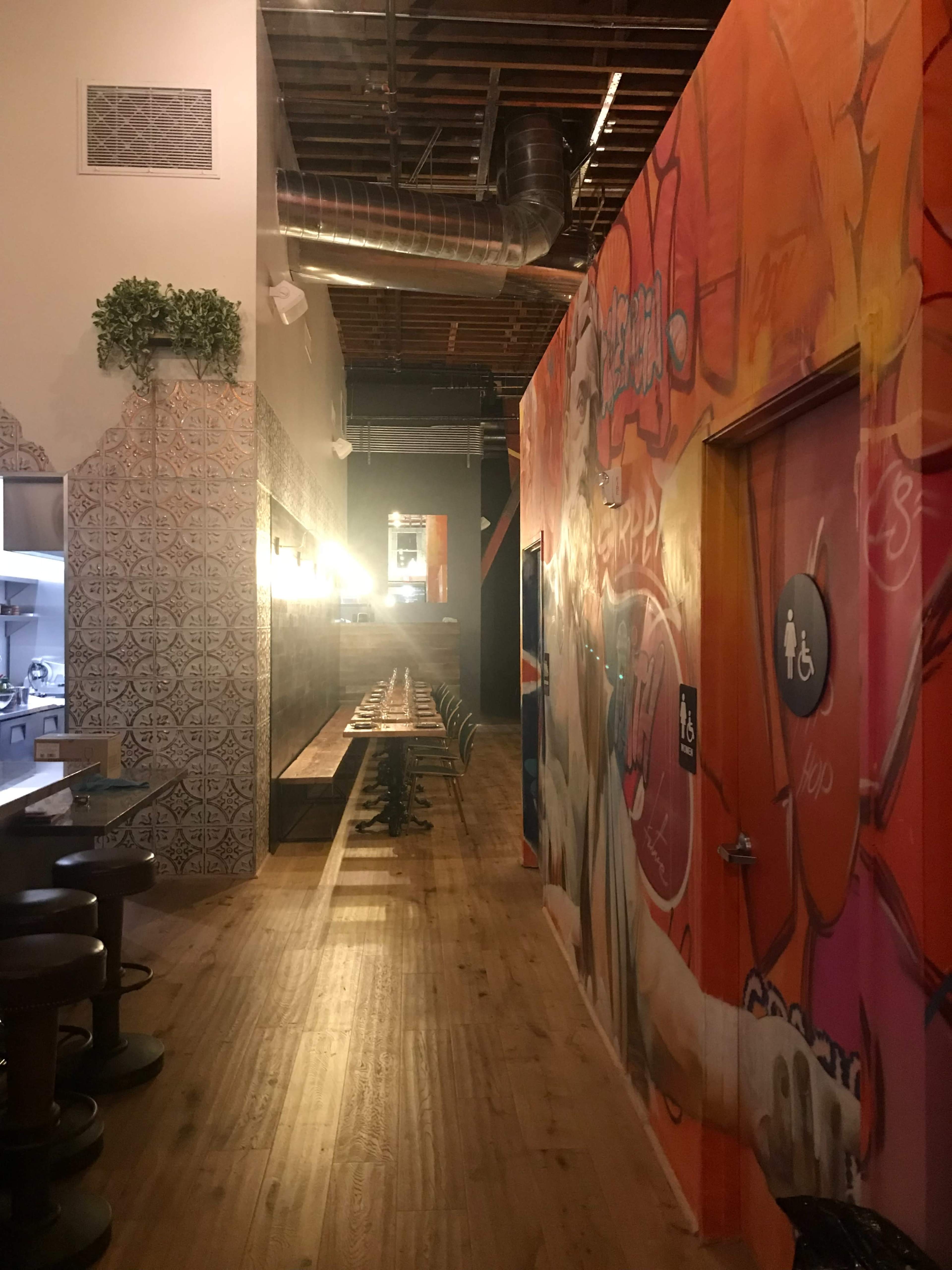 The image shows a narrow hallway in a restaurant, featuring an orange and pink graffiti-covered wall, with a long wooden table on one side and a kitchen area visible at the end.