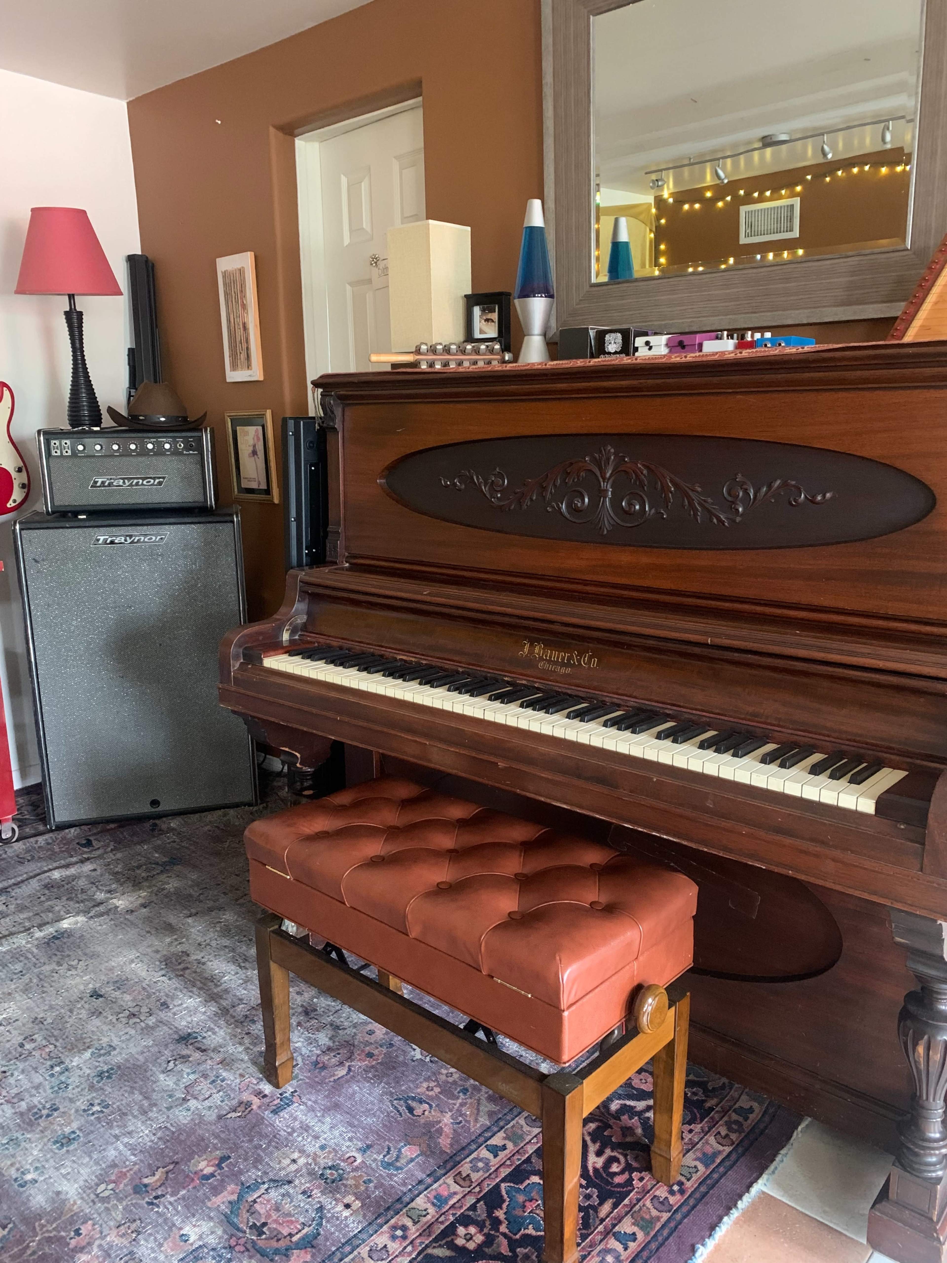 A wooden piano with a tufted leather bench in front of it, situated next to an amplifier and a decorative mirror.